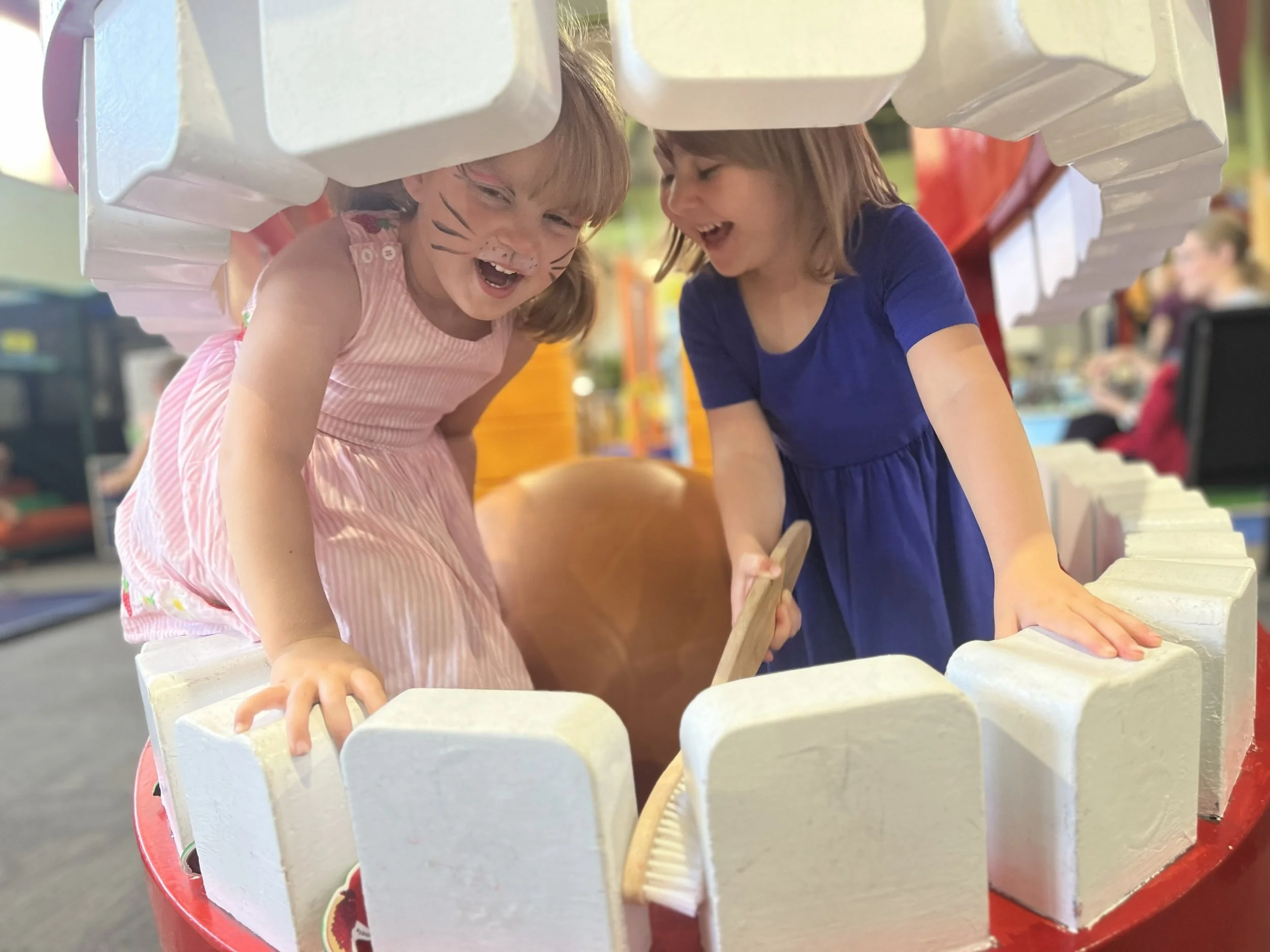 Two young girls with face paint playing a game with a wooden mallet and white rectangular blocks at an outdoor fair or carnival.