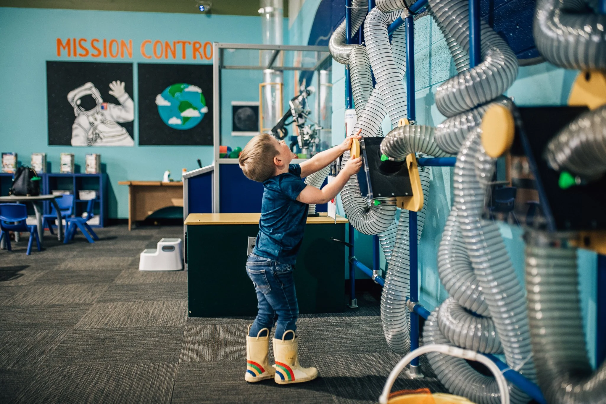 A young boy in yellow rain boots and rainbow-striped socks is reaching towards a control panel with large, flexible silver tubes attached to the wall in a science museum or educational space. The background features a mural with an astronaut, Earth, and the text 'MISSION CONTROL'.