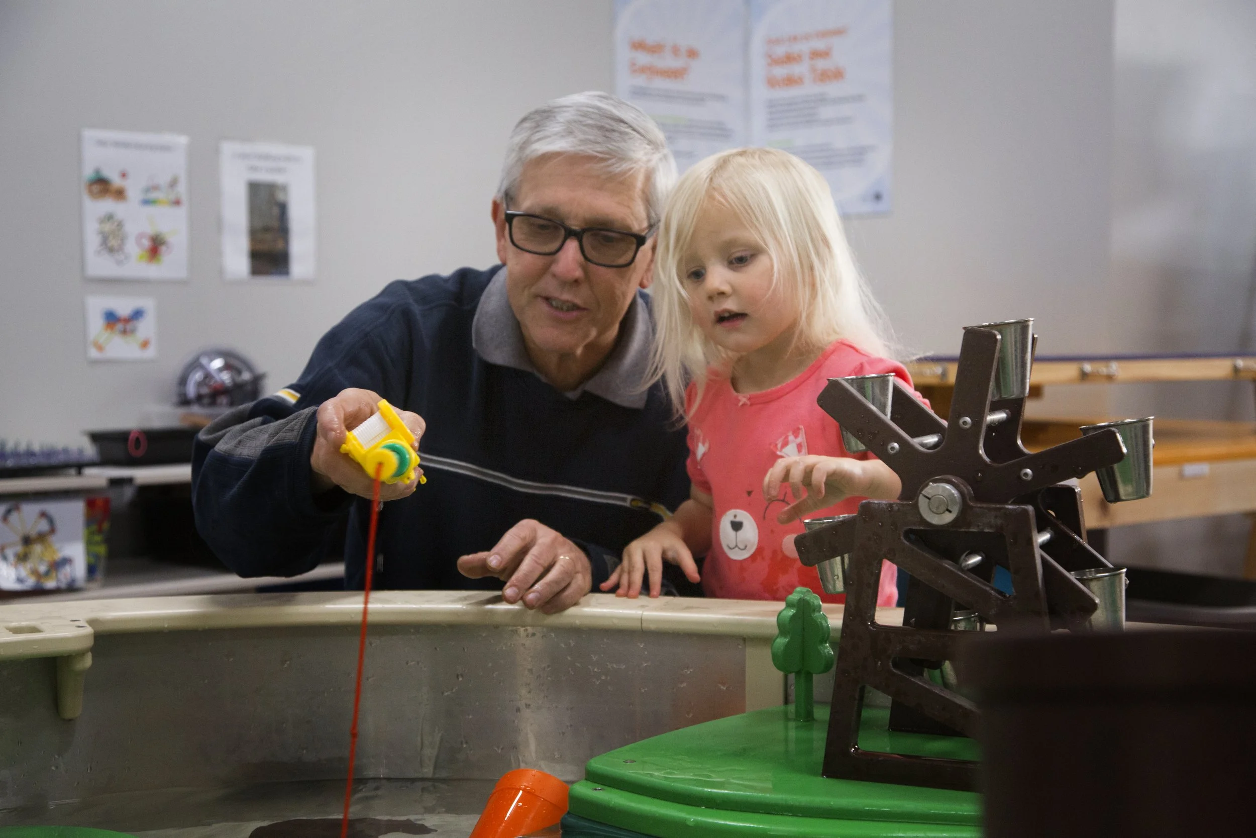 An elderly man and a young girl are engaged in a science or engineering activity at a table, examining a small yellow robot. The setting appears to be a classroom or activity room, with educational posters and toys in the background.