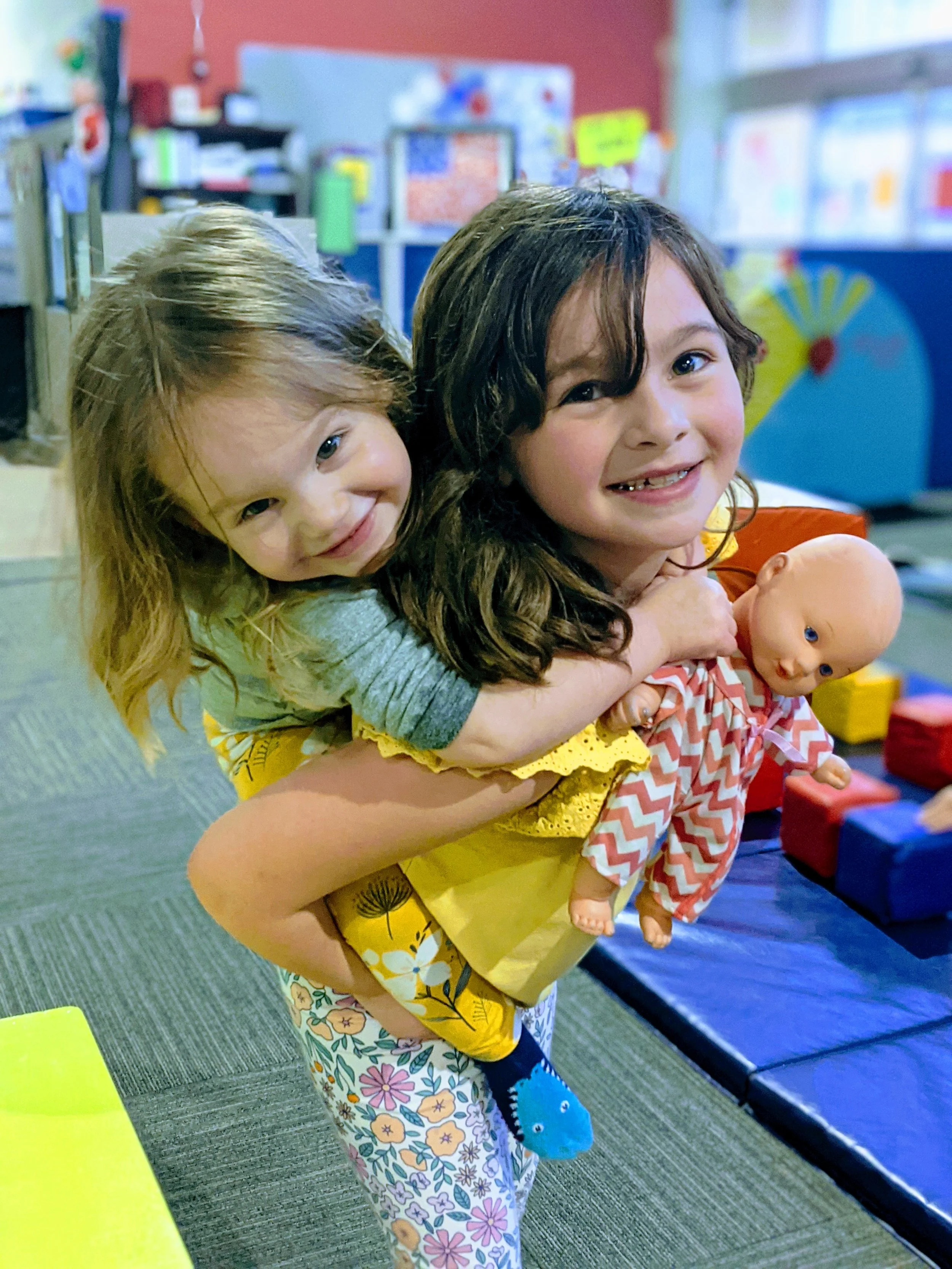 Two young girls, one with light brown hair and the other with dark brown hair, holding a doll with blue eyes, in a colorful classroom with toys and educational materials in the background.