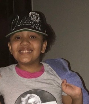 Young girl smiling, wearing a black Oakland Raiders cap and a gray T-shirt with a printed image.
