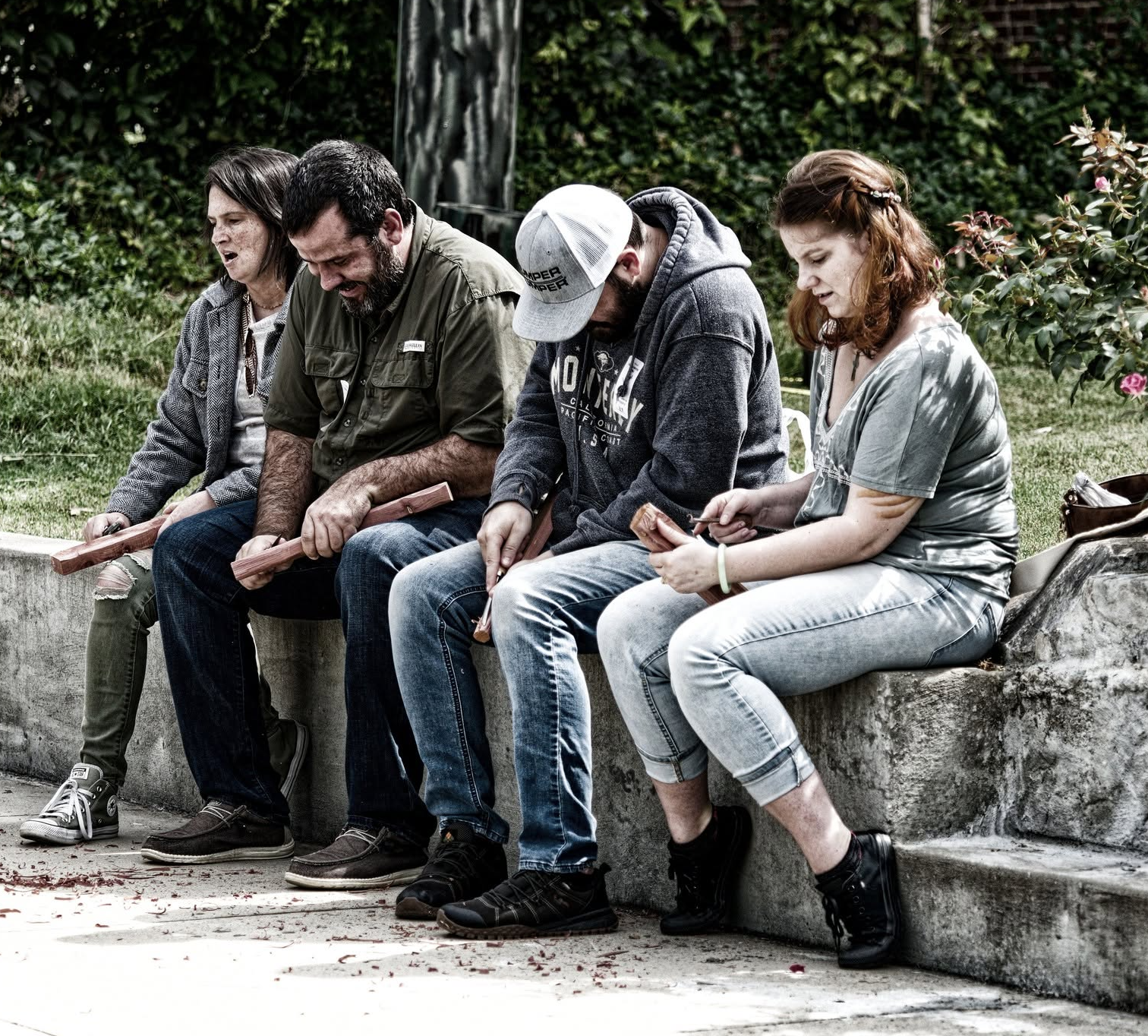 Four young adults sitting on a concrete bench outdoors, holding wooden blocks, appearing to joke or laugh with heads bowed.