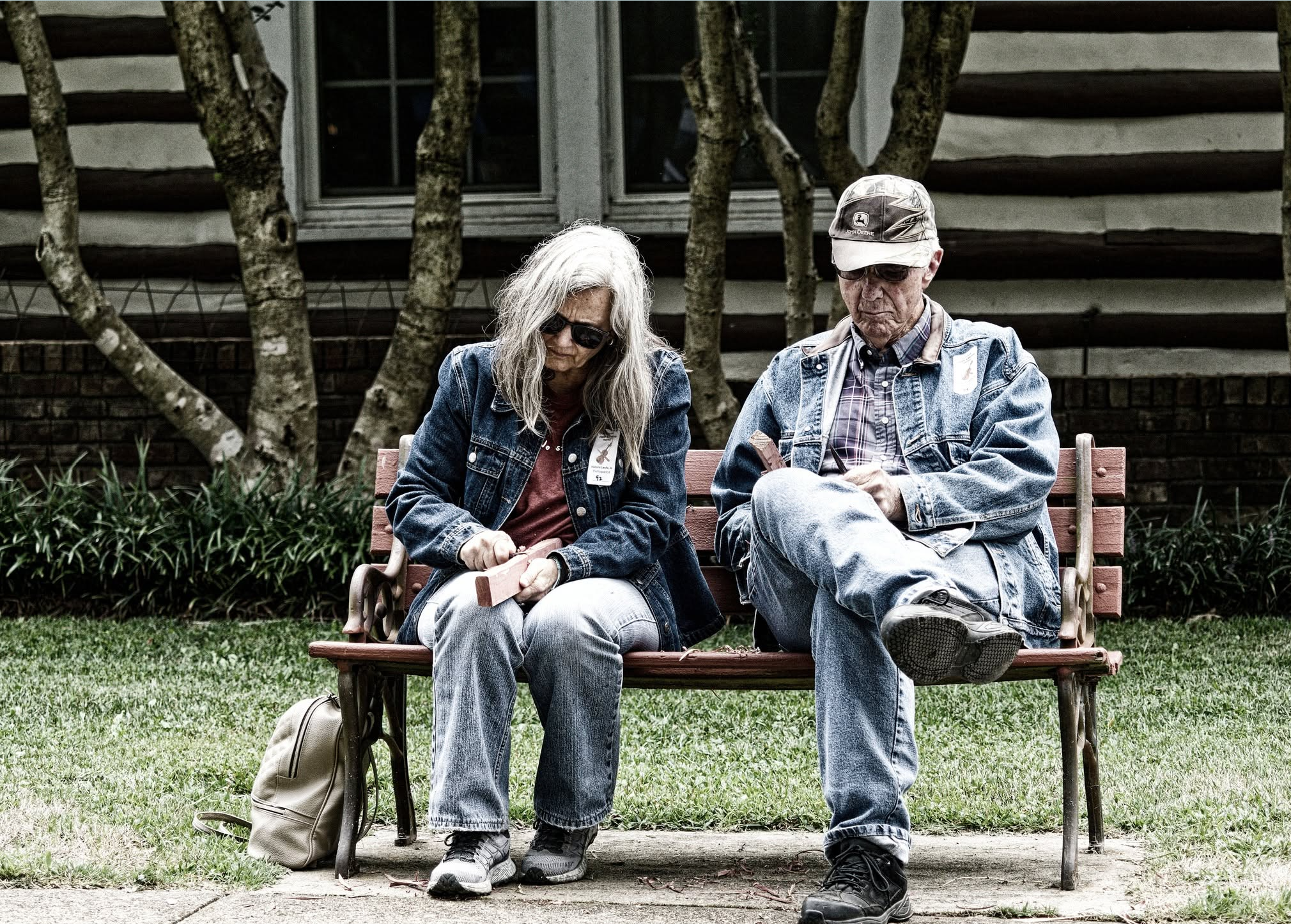 An older man and woman sitting on a park bench, looking at something in the woman's hand, with trees and a house in the background.