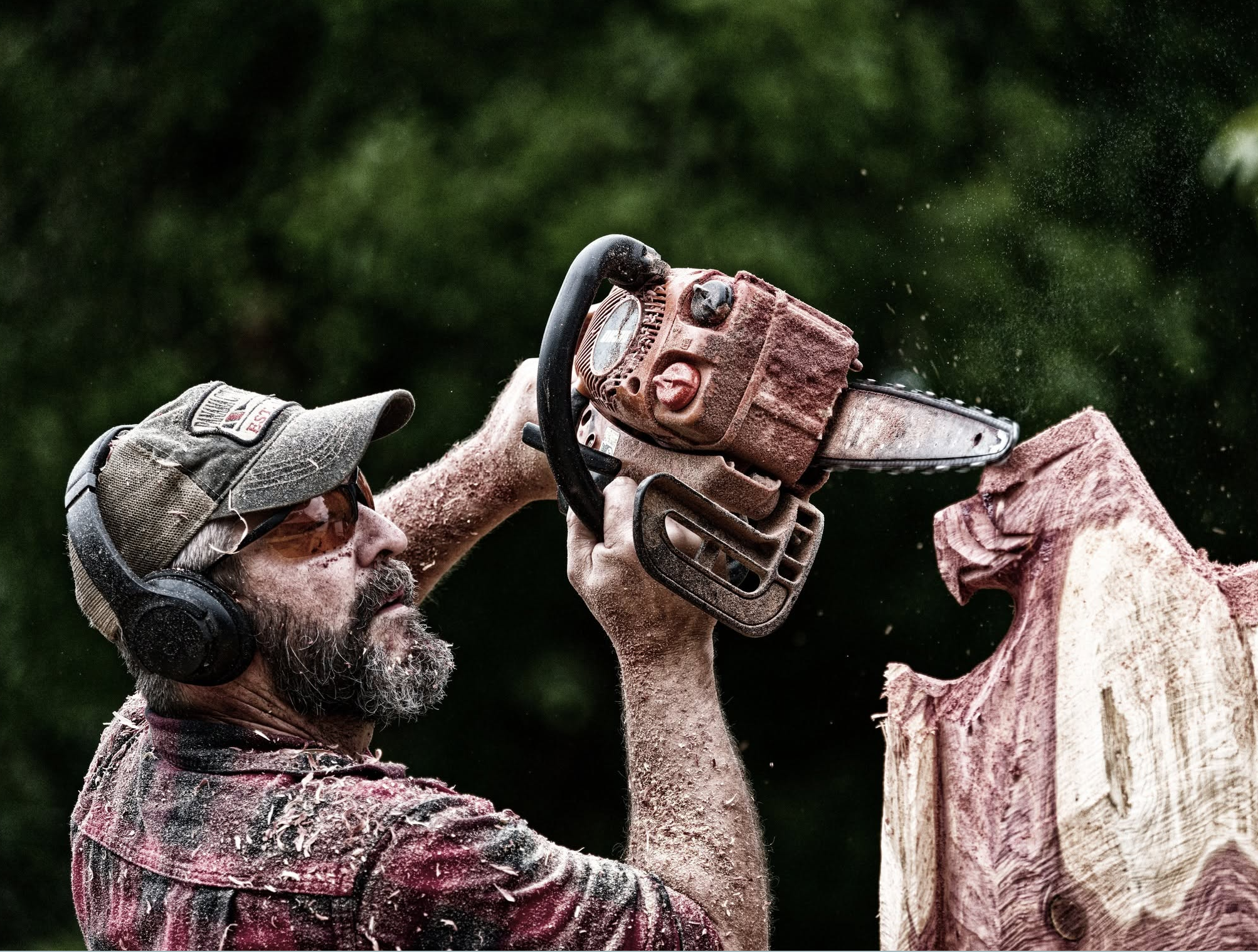 A man with a gray beard and sunglasses chiseling a piece of wood outdoors. Dust and wood particles are flying through the air. The man is wearing ear protection and a plaid shirt covered in sawdust. Another person is partially visible holding the wood steady.