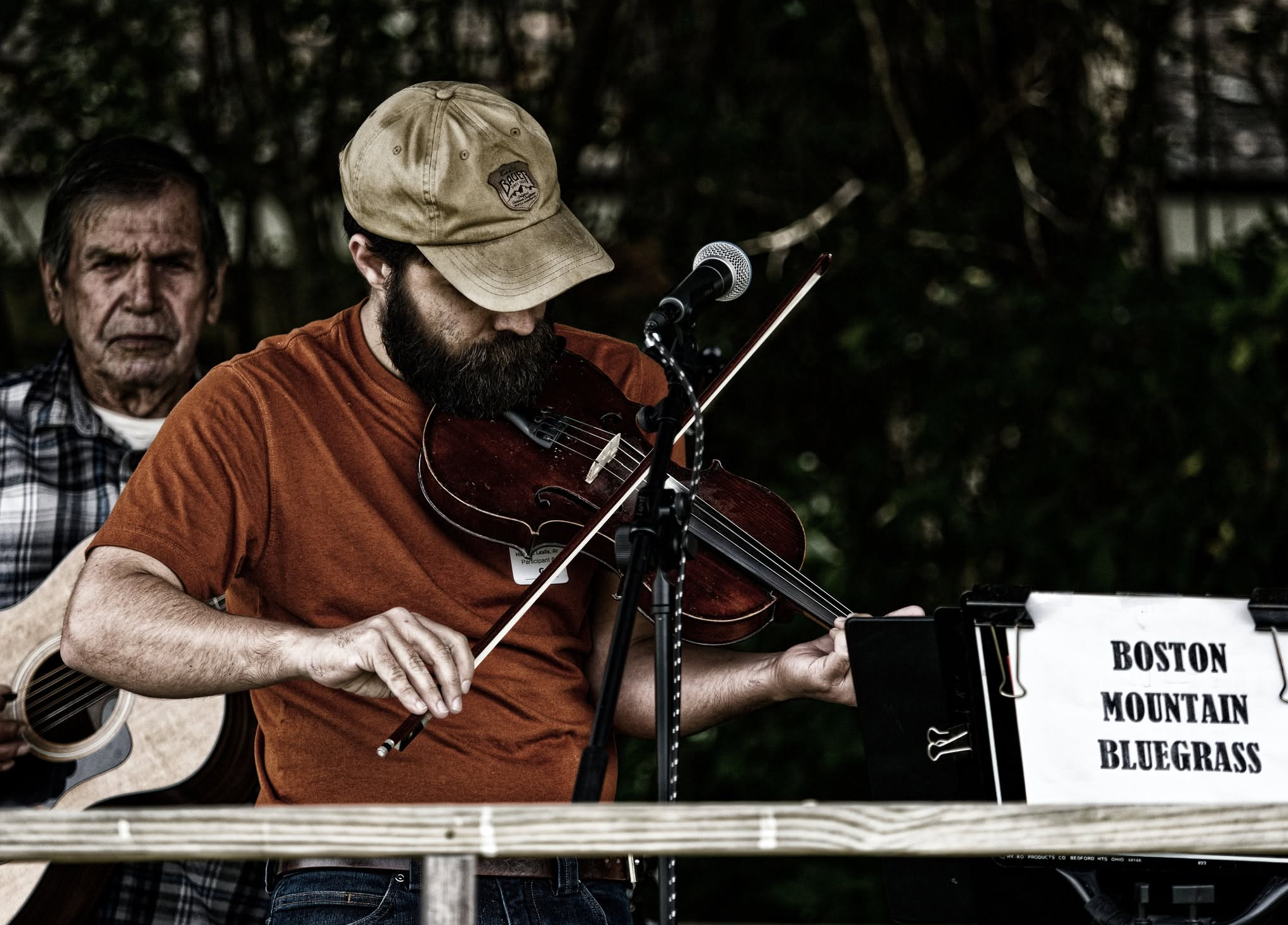 A man with a beard wearing a beige cap and orange t-shirt playing a violin during an outdoor event, with a woman in the background and a sign on the music stand that reads 'BOSTON MOUNTAIN BLUEGRASS'.