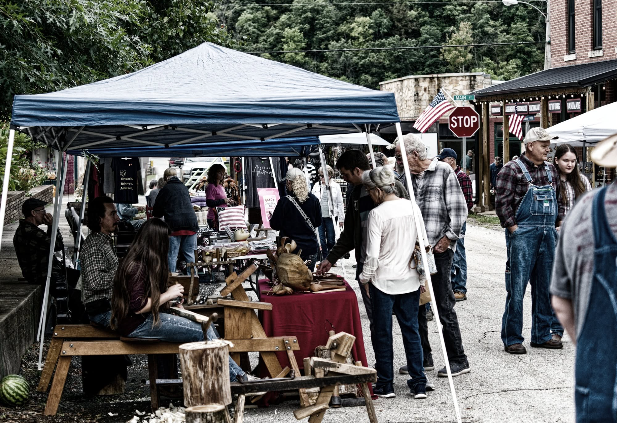A bustling outdoor market on a street corner, with several vendor booths under tents selling various items, and many people browsing and shopping. There are trees, a brick building, and a stop sign visible in the background.