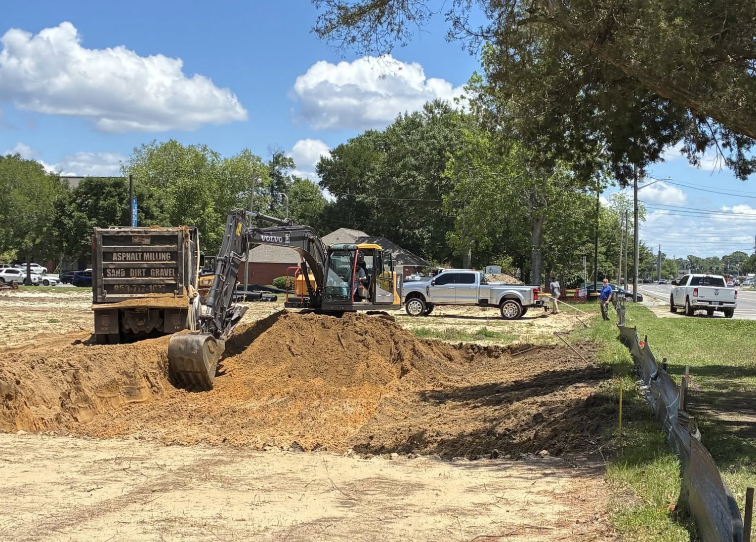 Construction site with an excavator digging a trench and a truck carrying dirt, surrounded by trees, vehicles, and a few workers.