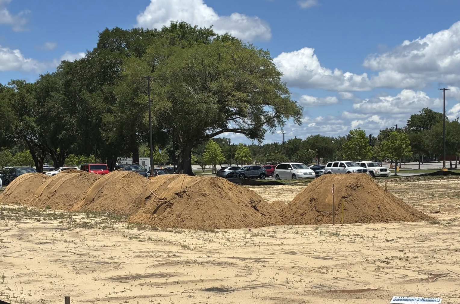 Four mounds of dirt on a cleared lot with a parking lot and trees in the background under a partly cloudy sky.