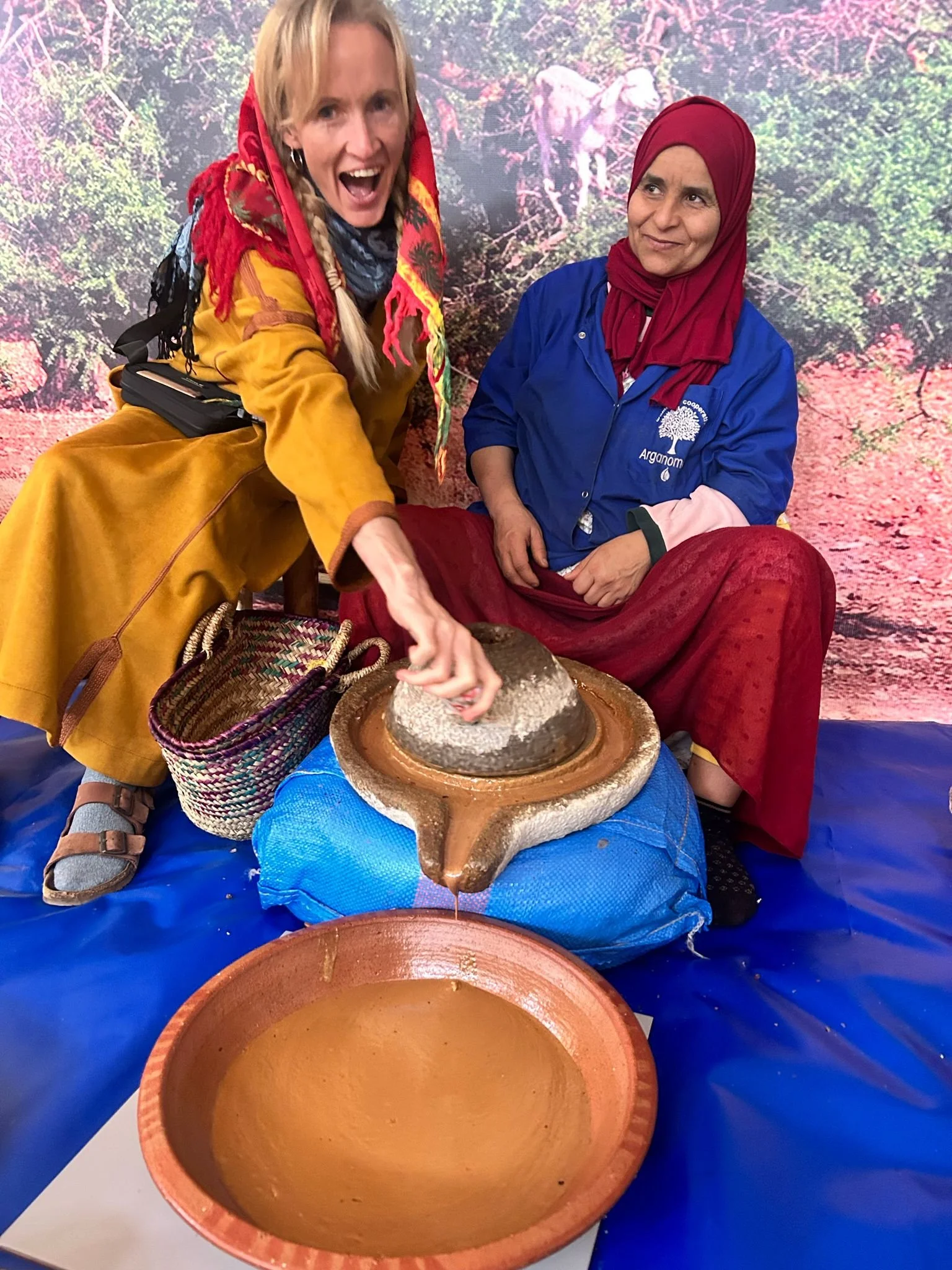 Two women, one in colorful attire and the other wearing a blue coat and a red headscarf, are seated together in front of a background depicting a jungle scene. The woman in the colorful outfit is smiling widely while the other looks at her with a neutral expression. They are working with clay on a table, shaping it in a pottery-making activity.