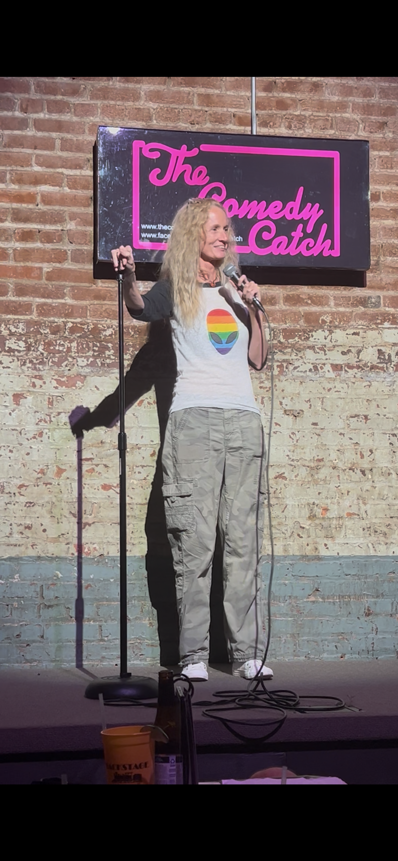 A comedian on stage at The Comedy Catch, holding a microphone in front of a brick wall and a neon sign. She is wearing a rainbow-colored LGBT pride heart t-shirt and beige pants.