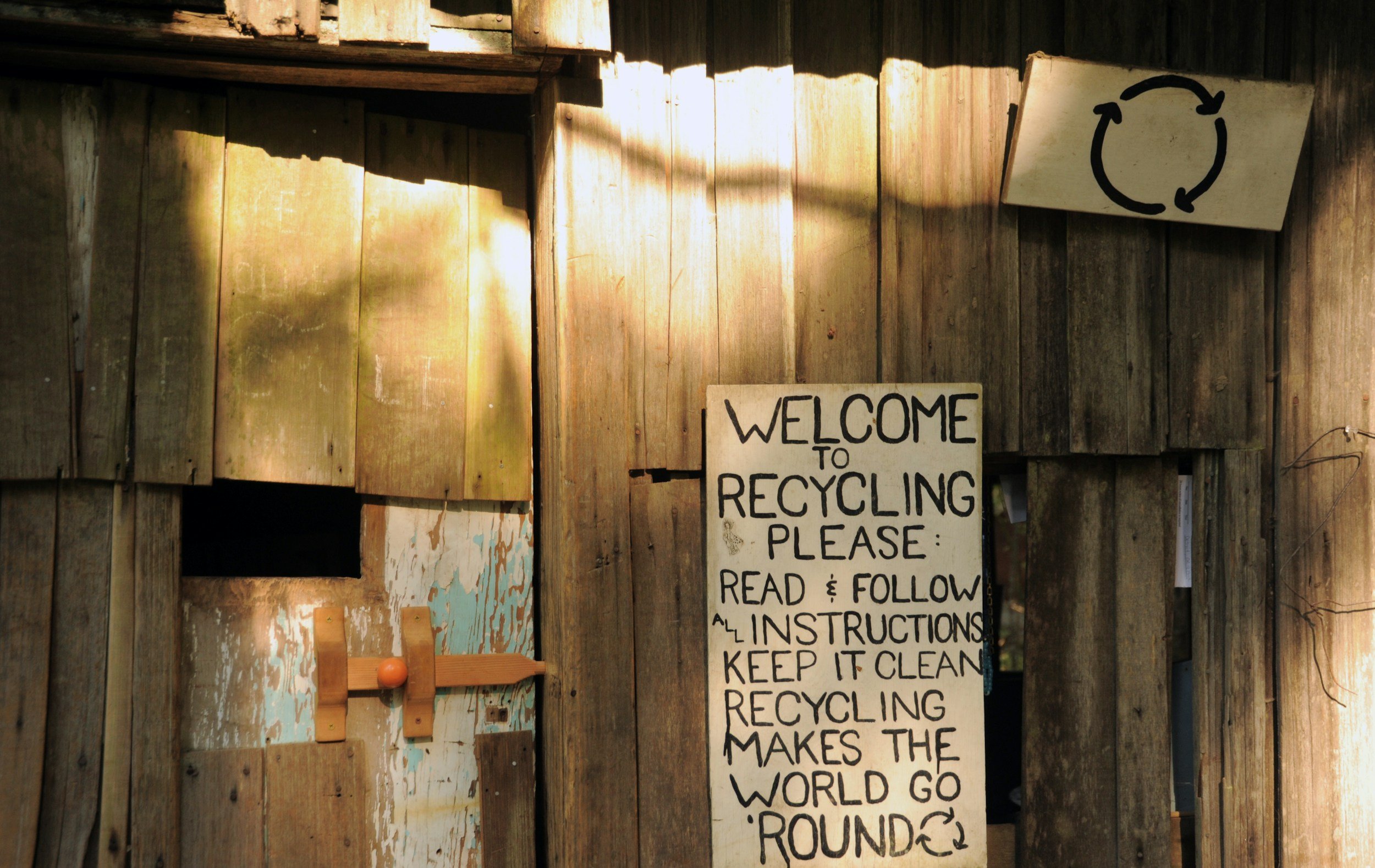 A wooden wall with a recycling sign and a handwritten note that says 'Welcome to recycling. Please read & follow all instructions. Keep it clean. Recycle makes the world go round.' There is also a smaller sign with a recycling symbol on it.