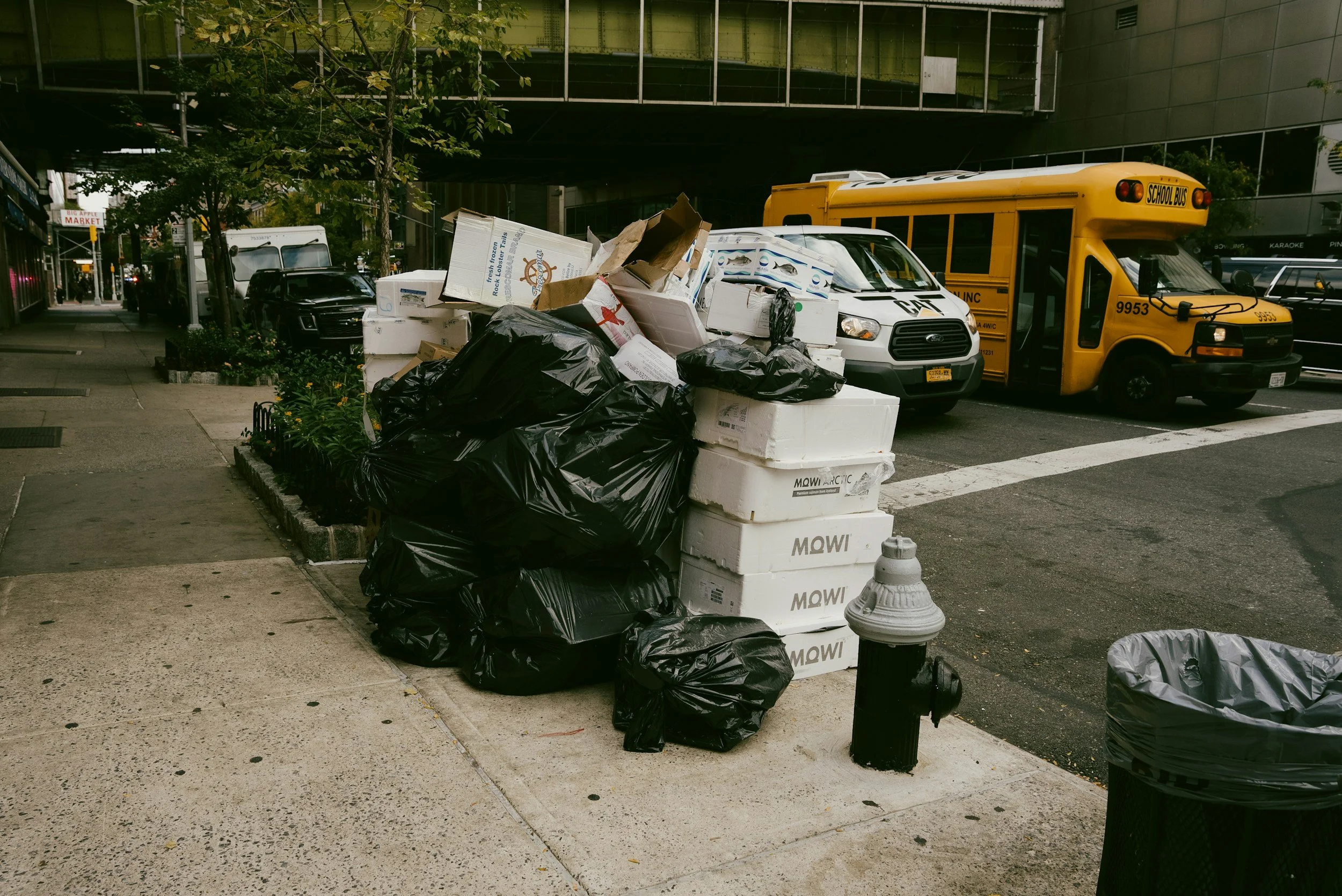 Street scene with garbage bags, cardboard boxes, and Styrofoam containers piled on the sidewalk near parked cars and a yellow school bus.