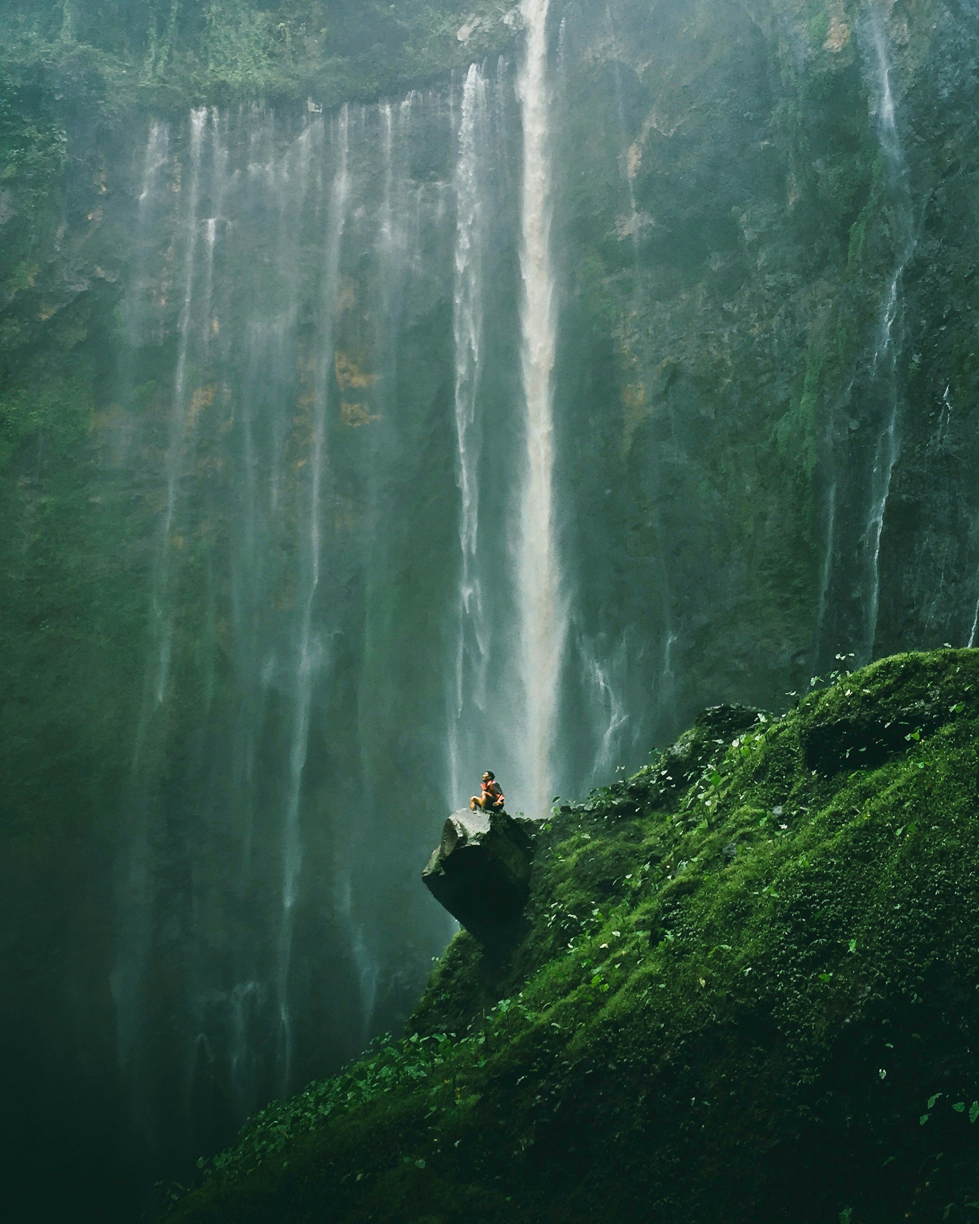 A person sitting on a rock beneath a tall waterfall in a lush green forest.