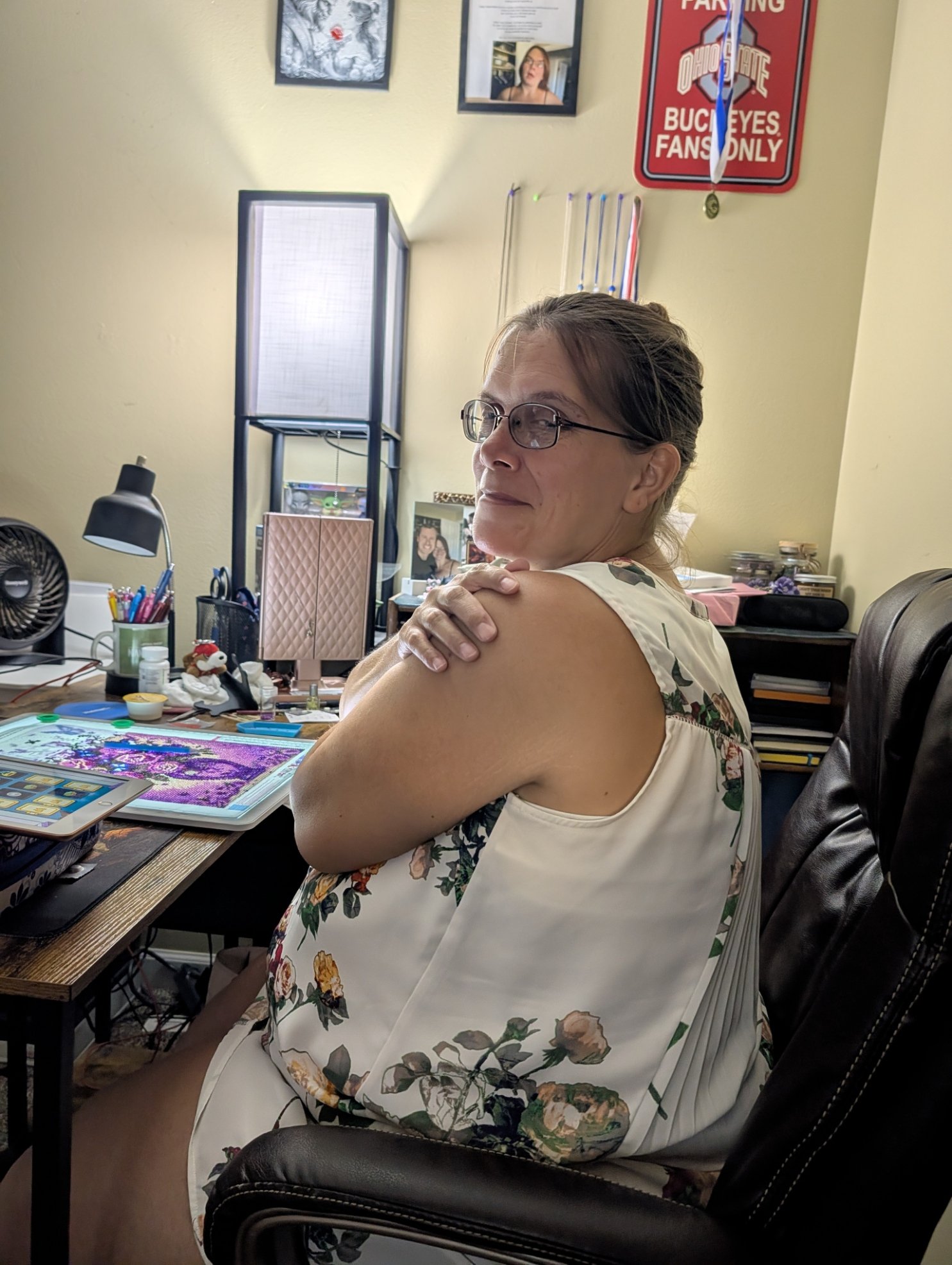 A woman with glasses and a floral dress sitting in a black office chair at a cluttered desk in a home office, with her arms crossed over her shoulders, looking slightly to her right, with framed photos and decorations on the wall behind her.