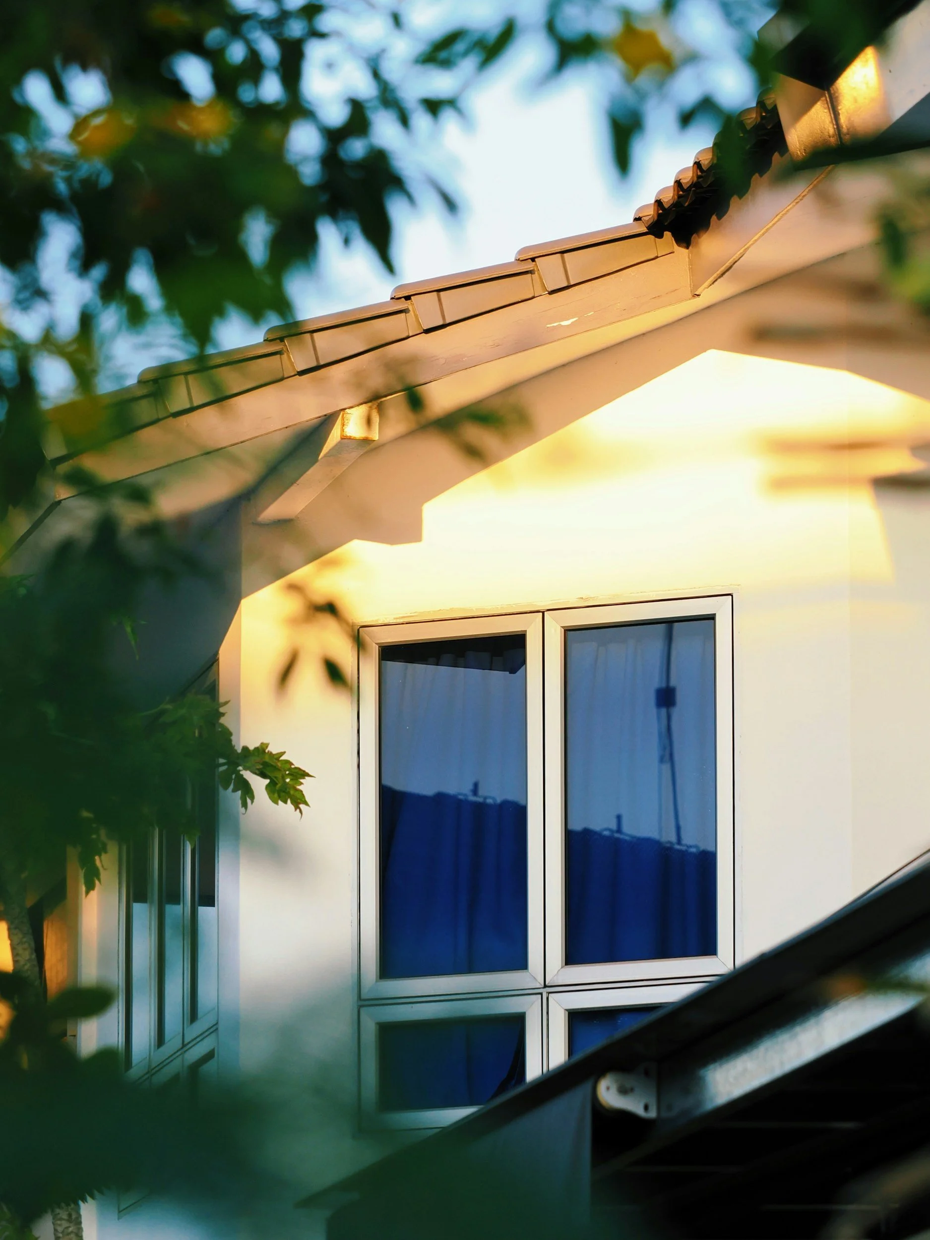 Photo of a house with a yellow exterior wall, large window with blue curtains, and a sloped roof with brown tiles, during sunset.