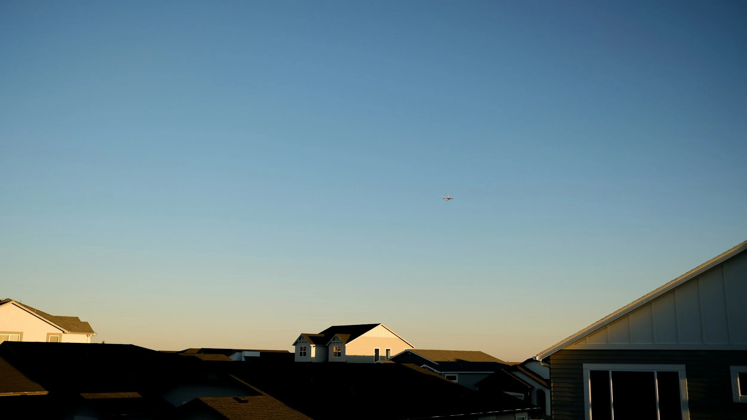 Clear blue sky with a small airplane flying above residential houses with peaked roofs, some windows visible, sunlight casting shadows.