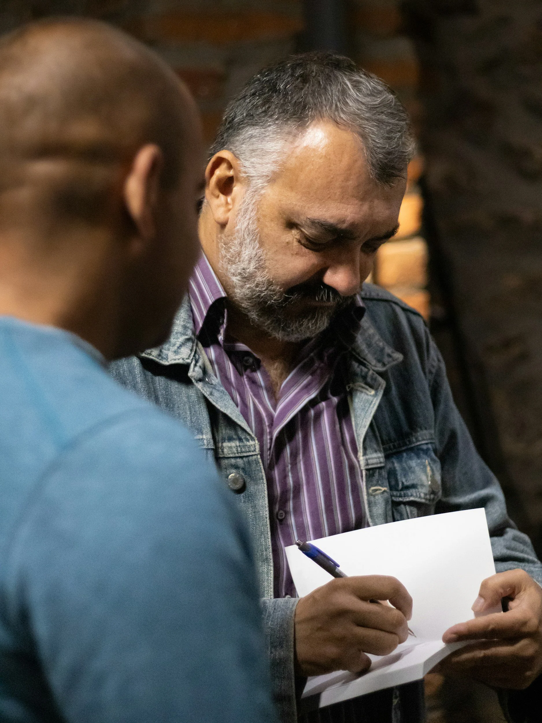 A man with gray hair and beard, wearing a striped purple shirt and denim jacket, writing on a piece of paper with a blue pen in a conversation setting.
