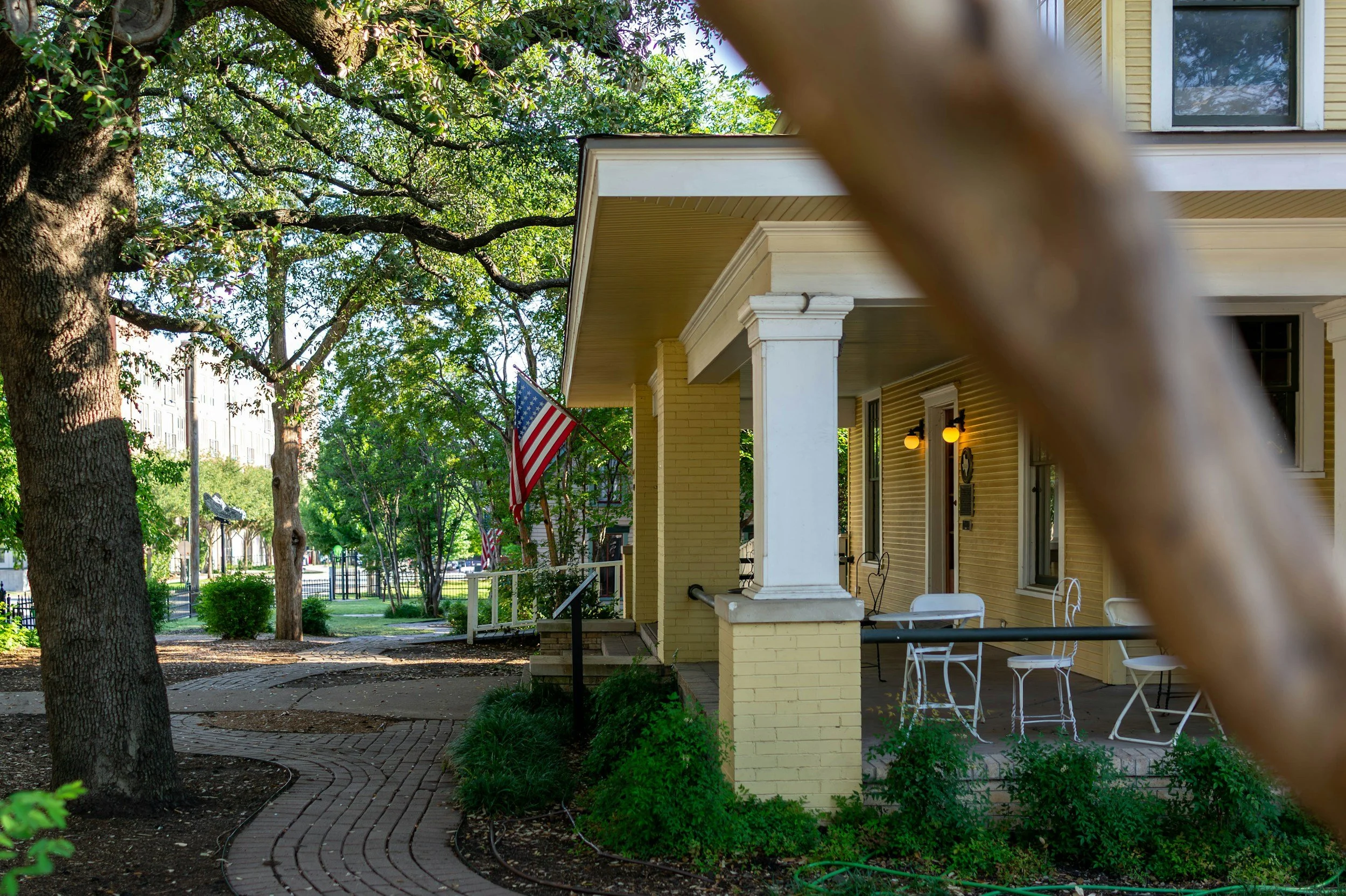 Side porch of a yellow house with white columns, chairs, and a table, shaded by trees, with an American flag hanging on the porch, and a tree branch partially obstructing the view.