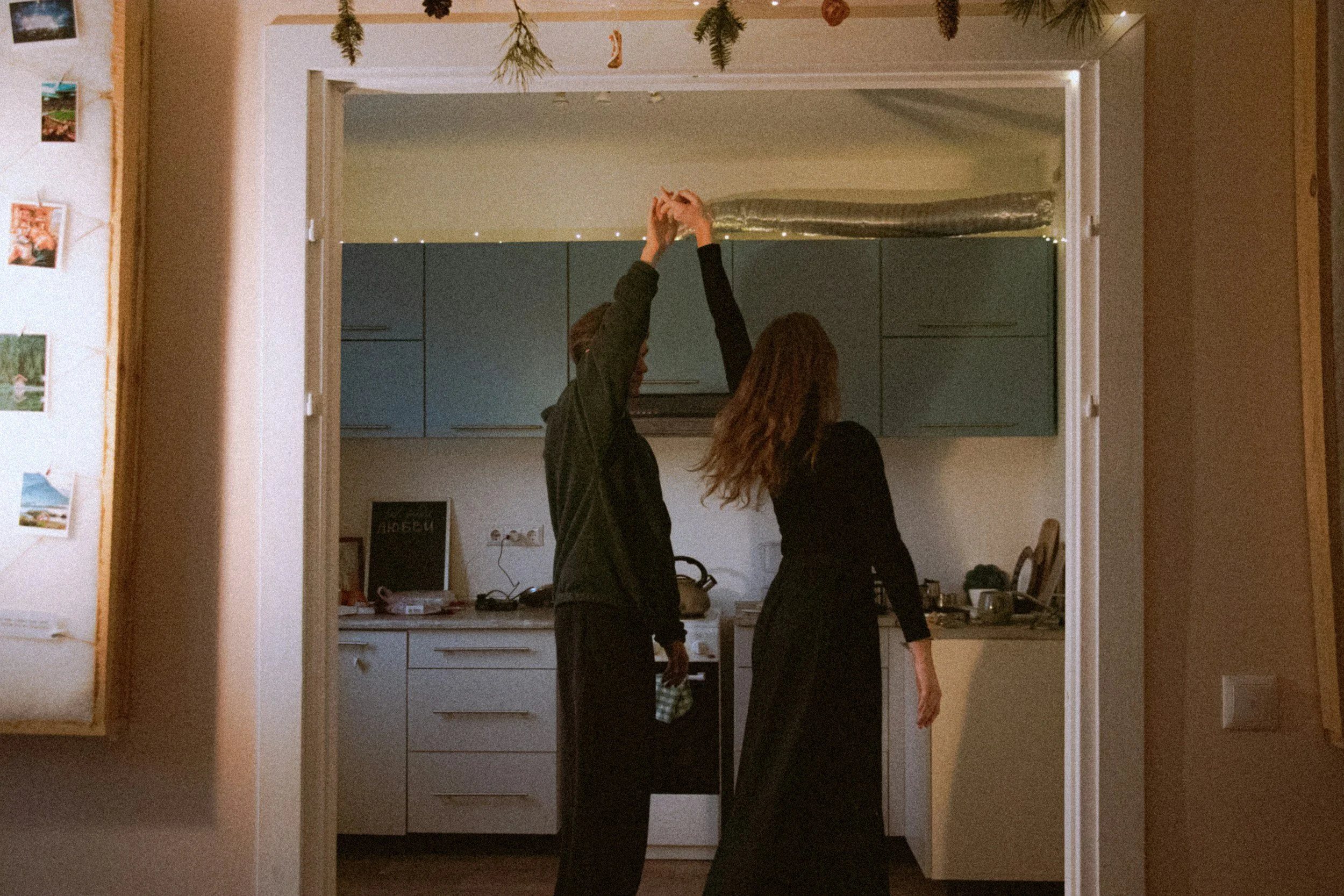 Two people are dancing or interacting in a kitchen, with one person raising their arm. The scene is viewed through a doorway, with a wall displaying small photos or postcards on the left side, and the kitchen has blue cabinets and some items on the counter.