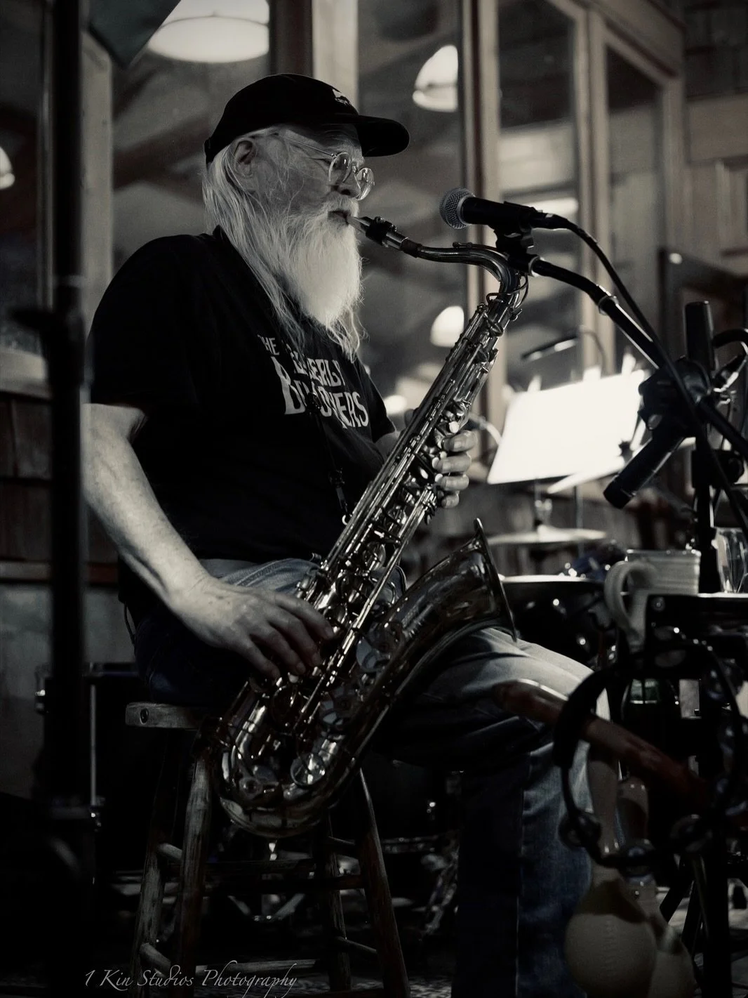 An elderly man with a long white beard, glasses, and a black cap playing a saxophone in a dimly lit indoor setting.