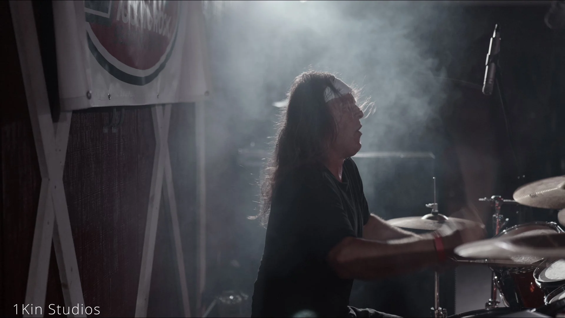 A male drummer playing drums in a dark, smoky environment with a bright light shining behind him.