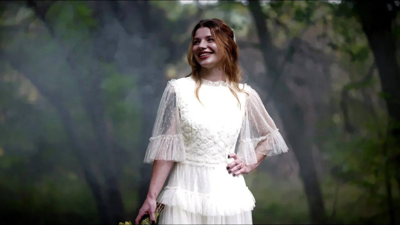 A woman with long red hair and red lipstick wearing a white lace dress with sheer, ruffled sleeves, standing outdoors in a wooded area with sunlight filtering through trees, smiling and holding a small bouquet.