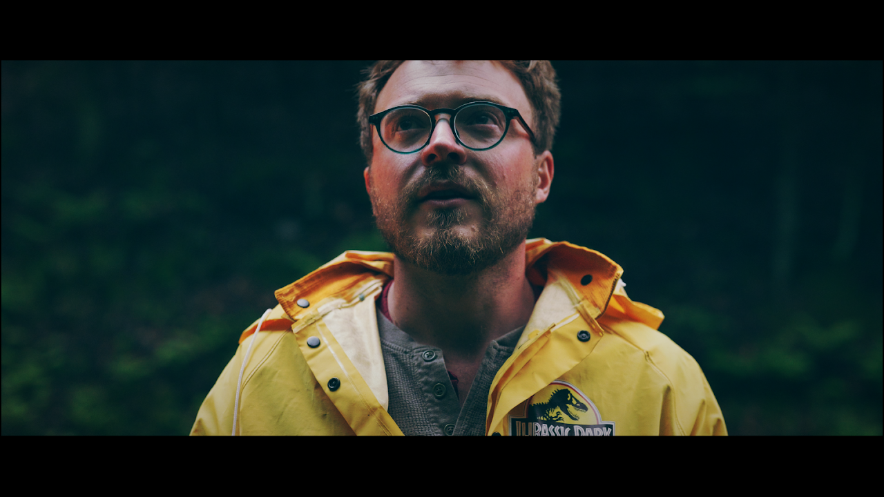 A man with glasses and a beard, wearing a yellow Jurassic Park jacket, standing outdoors with a blurred green forest background.
