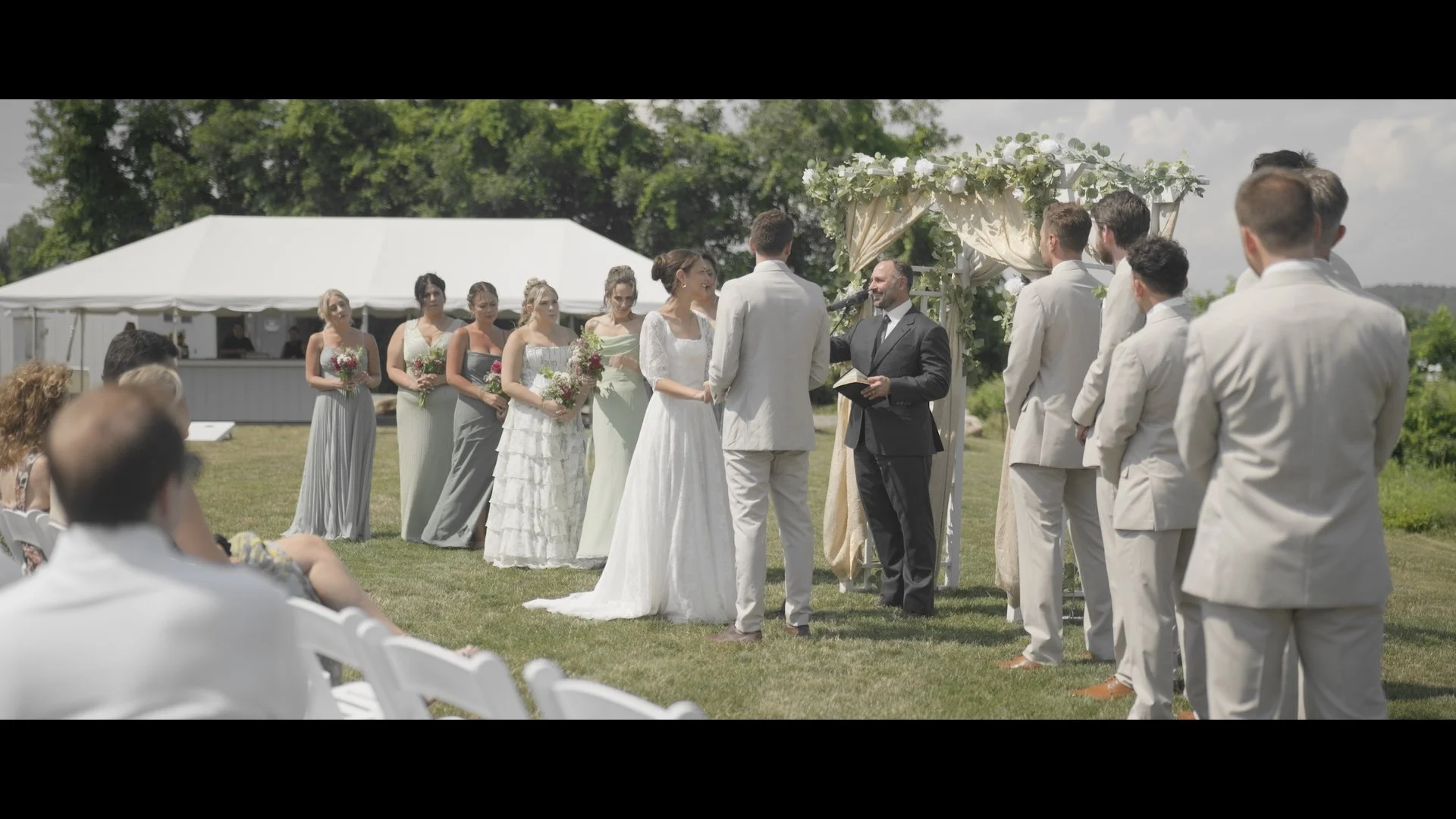 A couple getting married outdoors, with the officiant and wedding party standing in front of a decorated arch. Bridesmaids hold bouquets and guests seated in white chairs watch the ceremony.
