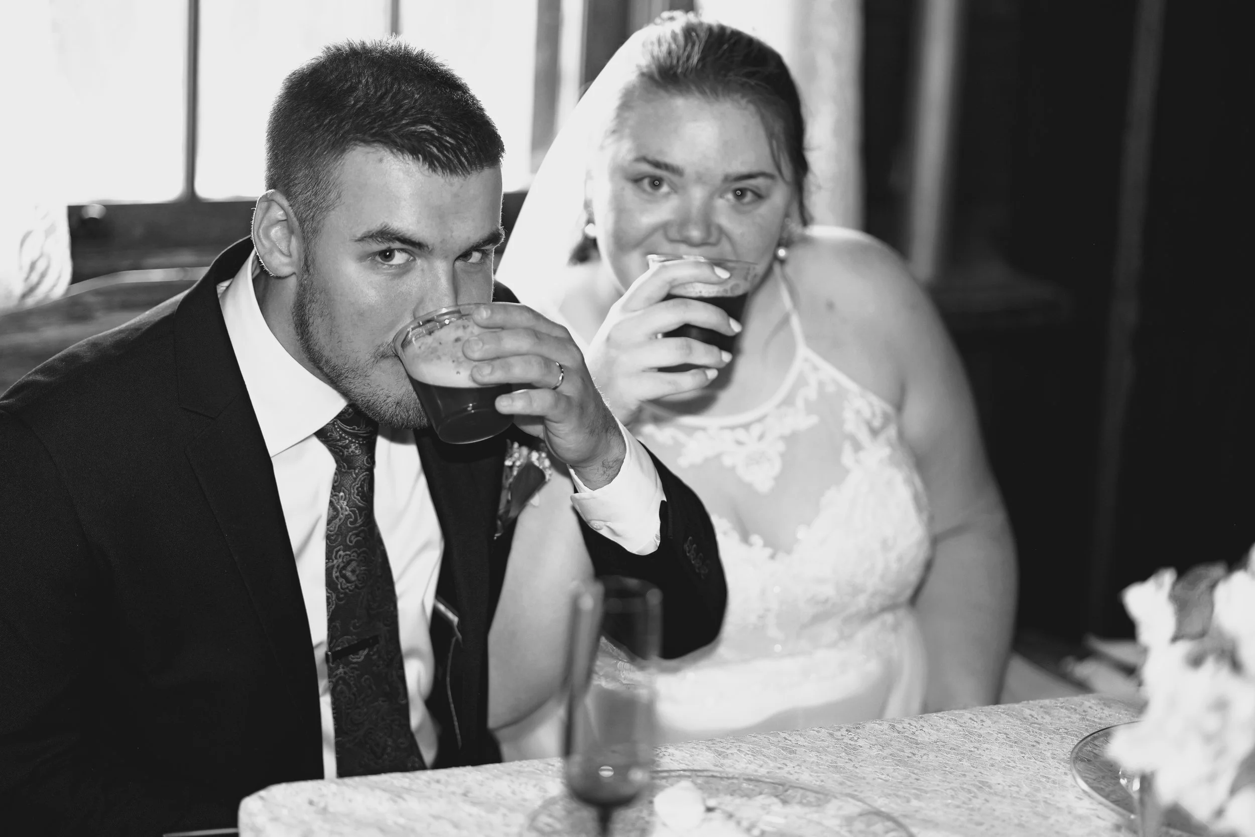 A black and white photo of a groom and bride drinking from glasses, seated at a table during a wedding reception.