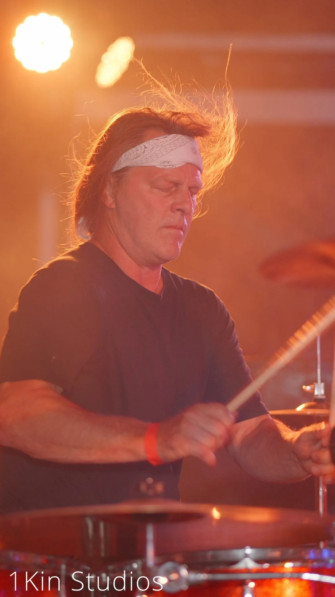 A man with long hair wearing a white bandana and a black t-shirt, playing the drums during a live music performance with warm stage lighting.