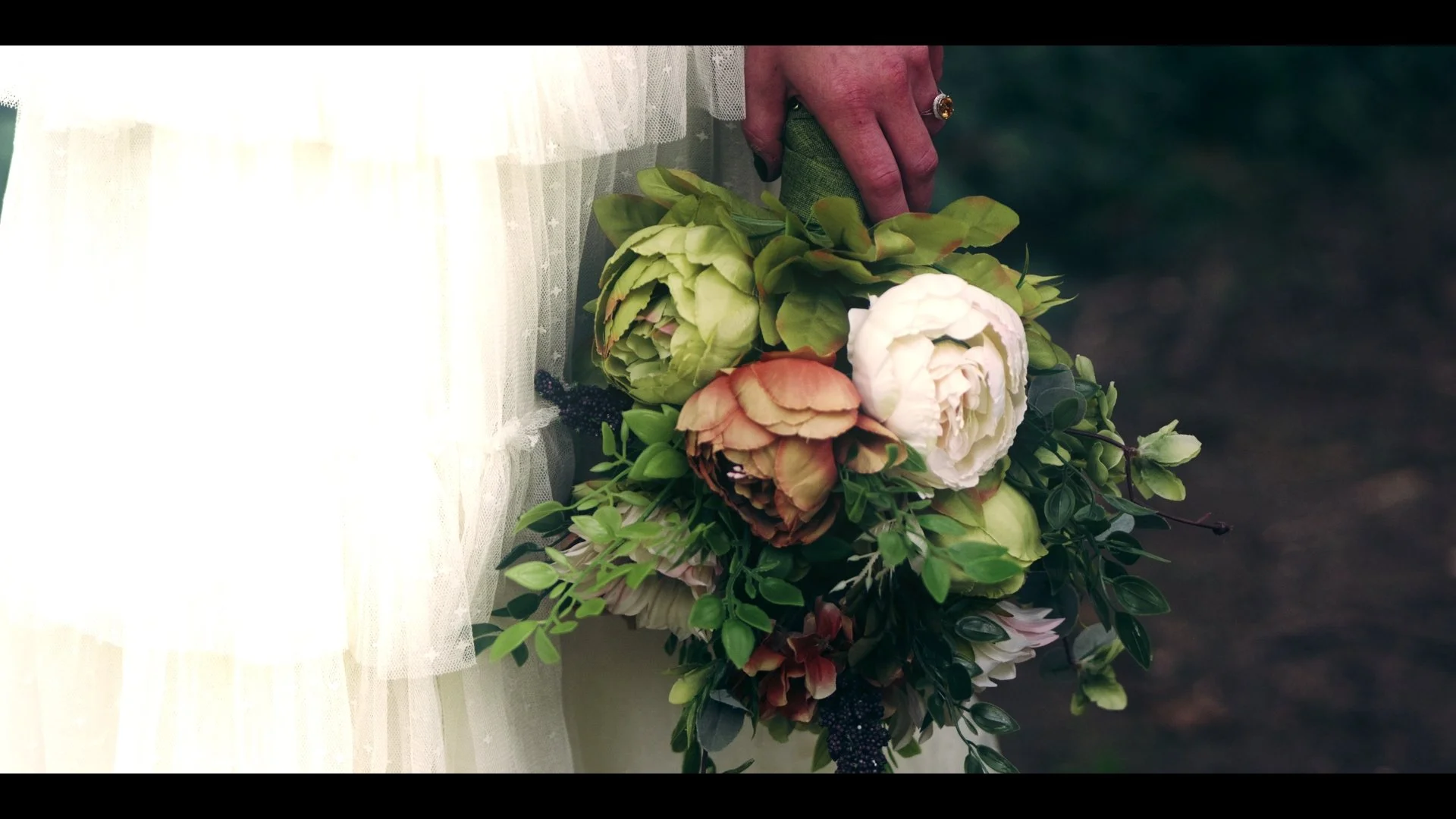 A person holding a bouquet of pink, white, and green flowers, with a white tulle skirt visible on the left side of the image and a dark blurred background.