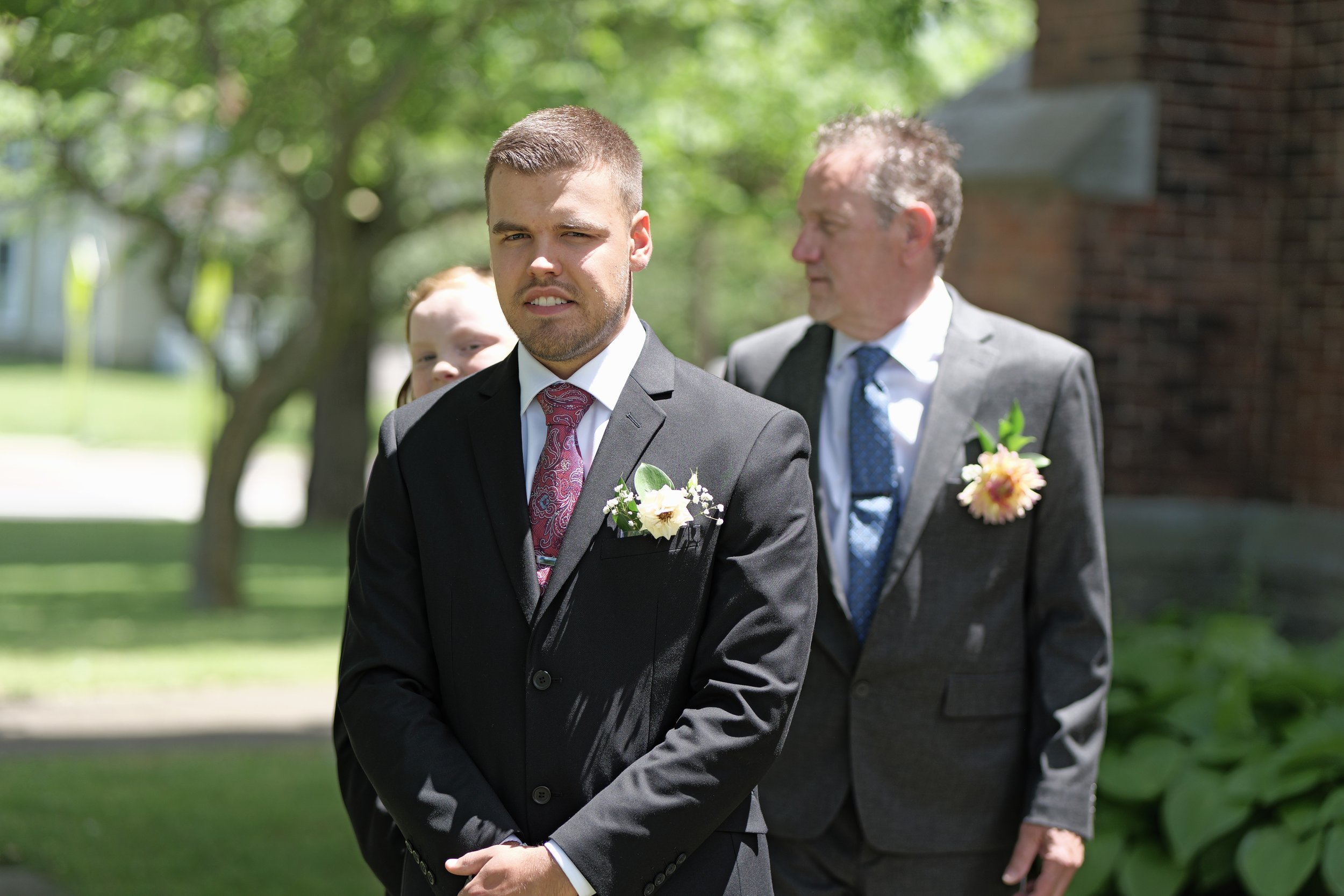 A young man in a black suit, white shirt, and red patterned tie standing outdoors during a sunny day. He has a flower boutonniere on his lapel, and is slightly squinting at the camera. Behind him, an older man and a woman are also dressed formally, l