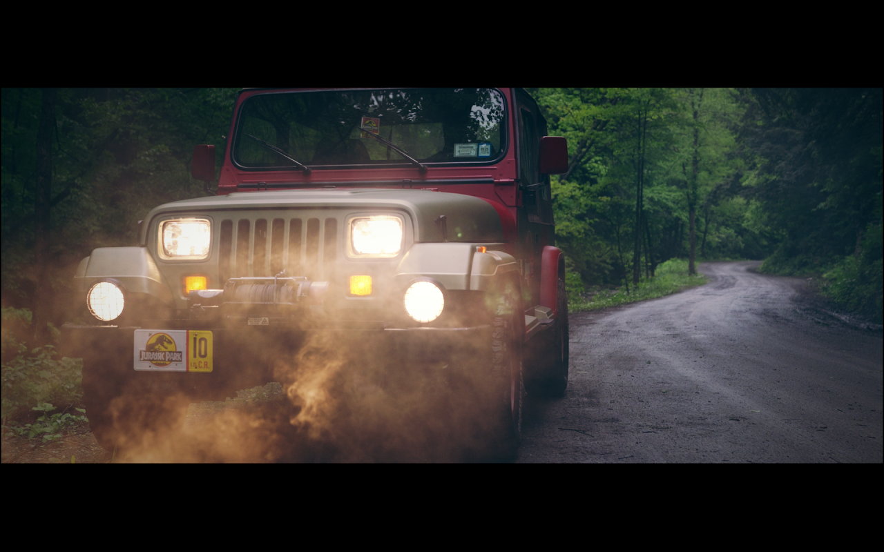 A red Jeep with its headlights on driving on a dirt road in a forest, kicking up dust.