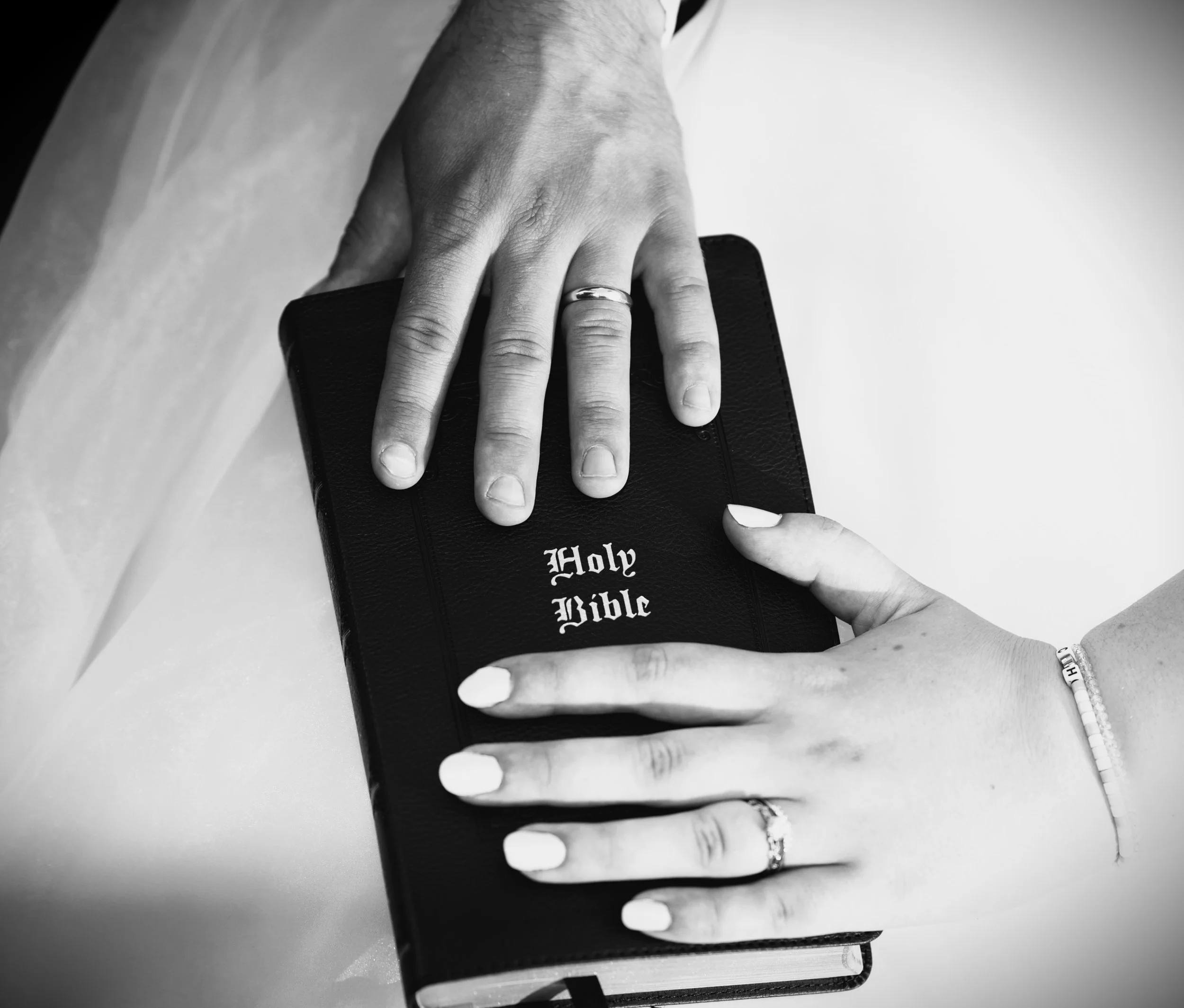 A hand with a wedding ring on the ring finger rests on a Bible labeled 'Holy Bible' on a table. A pen is positioned near the Bible.