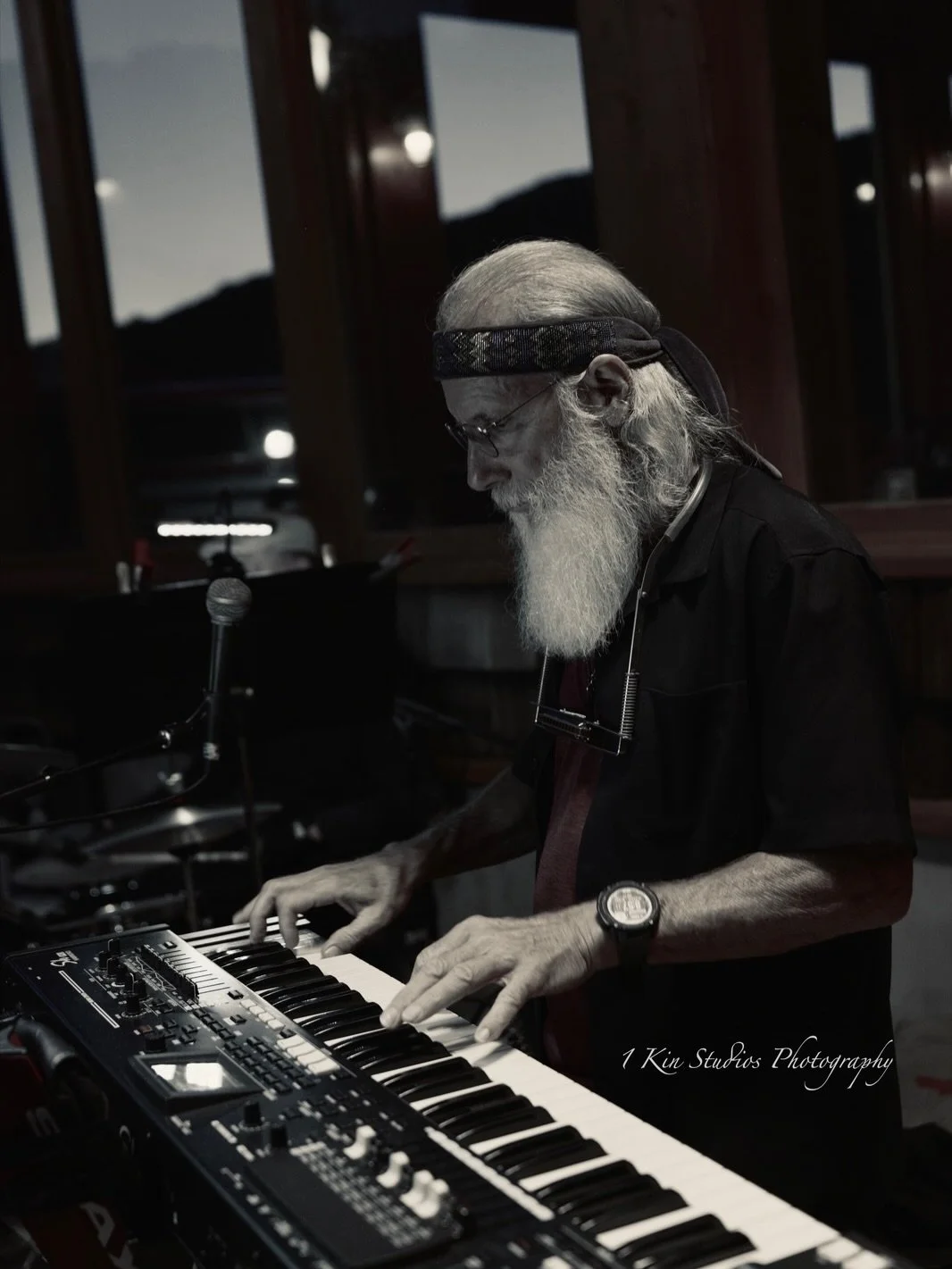 An elderly man with long white hair and beard playing a keyboard in a dimly lit room, wearing a black shirt, glasses, a headband, and a wristwatch, with a microphone nearby.