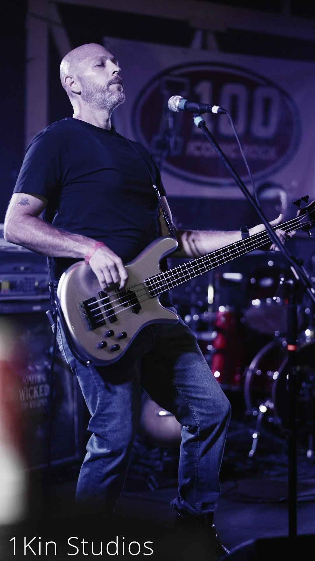 A male musician with a shaved head and beard playing a bass guitar on stage at a live music venue.
