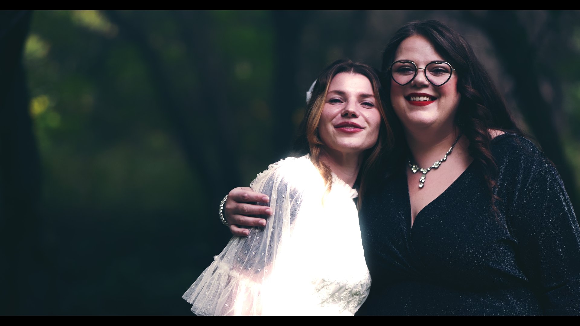 Two women embrace and smile outdoors, one wearing glasses and a dark dress with jewelry, the other in a white dress with sheer, polka-dotted sleeves.