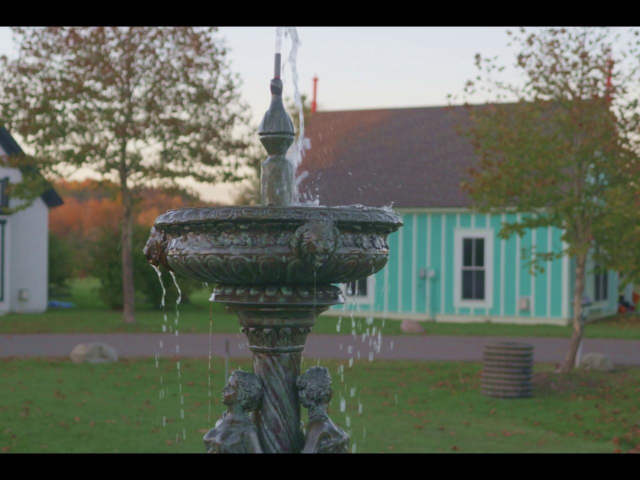 A decorative outdoor fountain with water flowing from the top, situated in a park or garden with trees and buildings in the background.