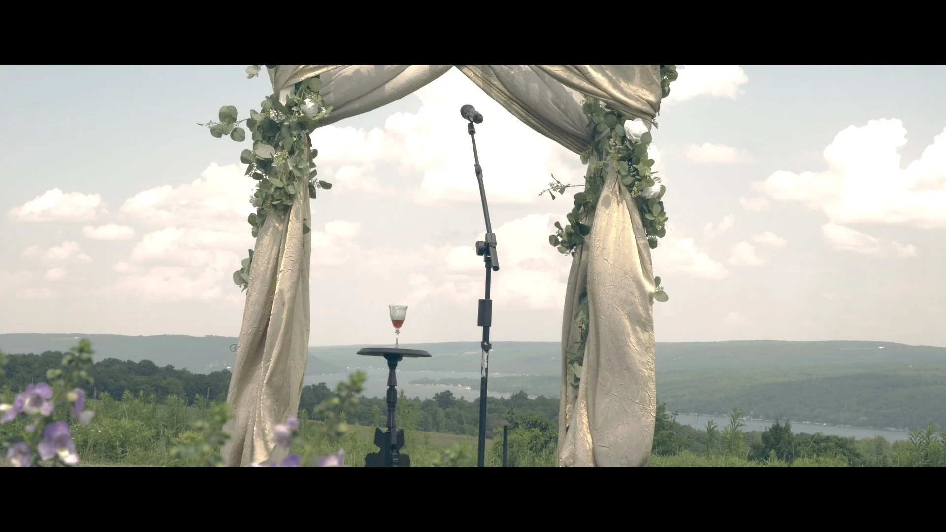 Outdoor wedding ceremony setup with a decorative draped arch and greenery, a microphone stand, a small table with a glass of drink, and a scenic landscape with water and hills in the background.