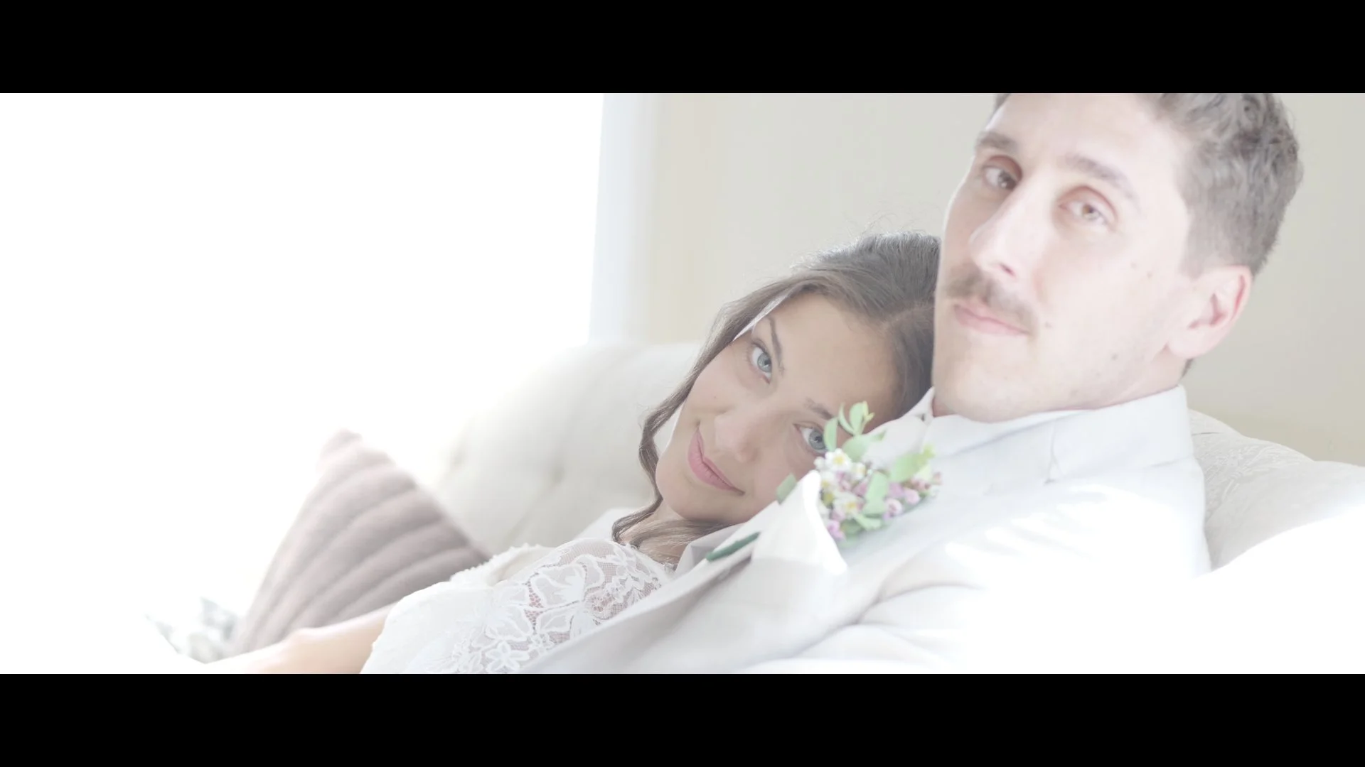 A bride and groom relaxing on a white sofa. The bride has her head on the groom's shoulder, and both are smiling softly. The bride is wearing a white lace dress, and the groom is in a white suit with a floral boutonniere.