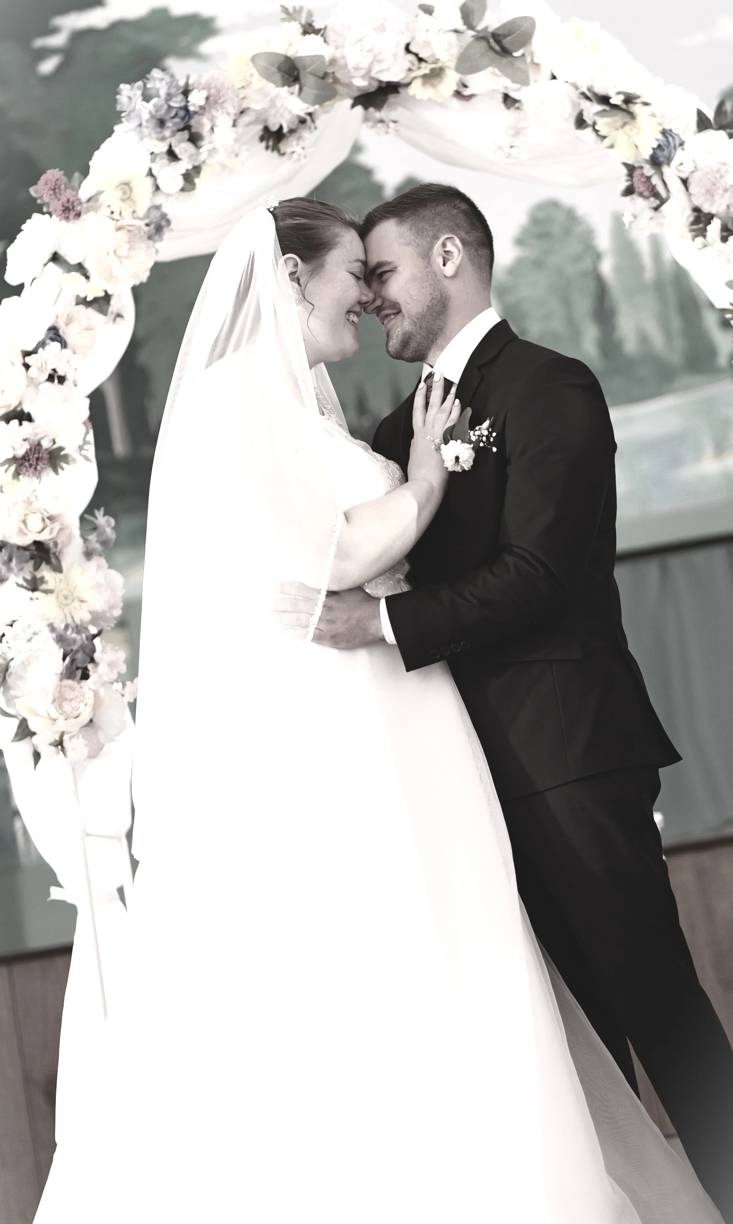 Bride and groom smiling and with foreheads touching beneath a floral arch on their wedding day.