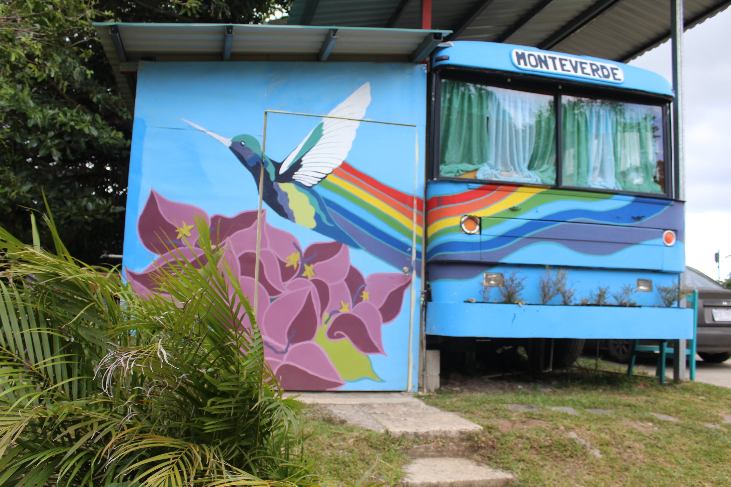 A colorful mural of a hummingbird with rainbow wings and a large pink flower on the side of a blue food truck labeled 'Monterde'. The truck is parked under a metal carport and surrounded by green plants.