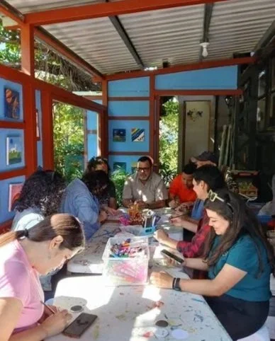 People sitting around a table engaging in arts and crafts activities outdoors, with blue walls and greenery in the background.