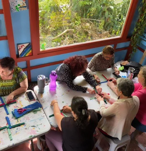 A group of women sitting around a table engaging in a craft activity, with supplies like scissors, paper, and water bottles, near a large window showing trees outside.