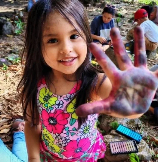 Young girl smiling and showing her painted hand during an outdoor art activity, with other children in the background sitting on the ground.