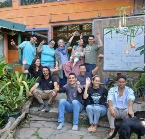 Group of nine people smiling and posing outdoors near a wooden building, with plants and a menu board in the background.