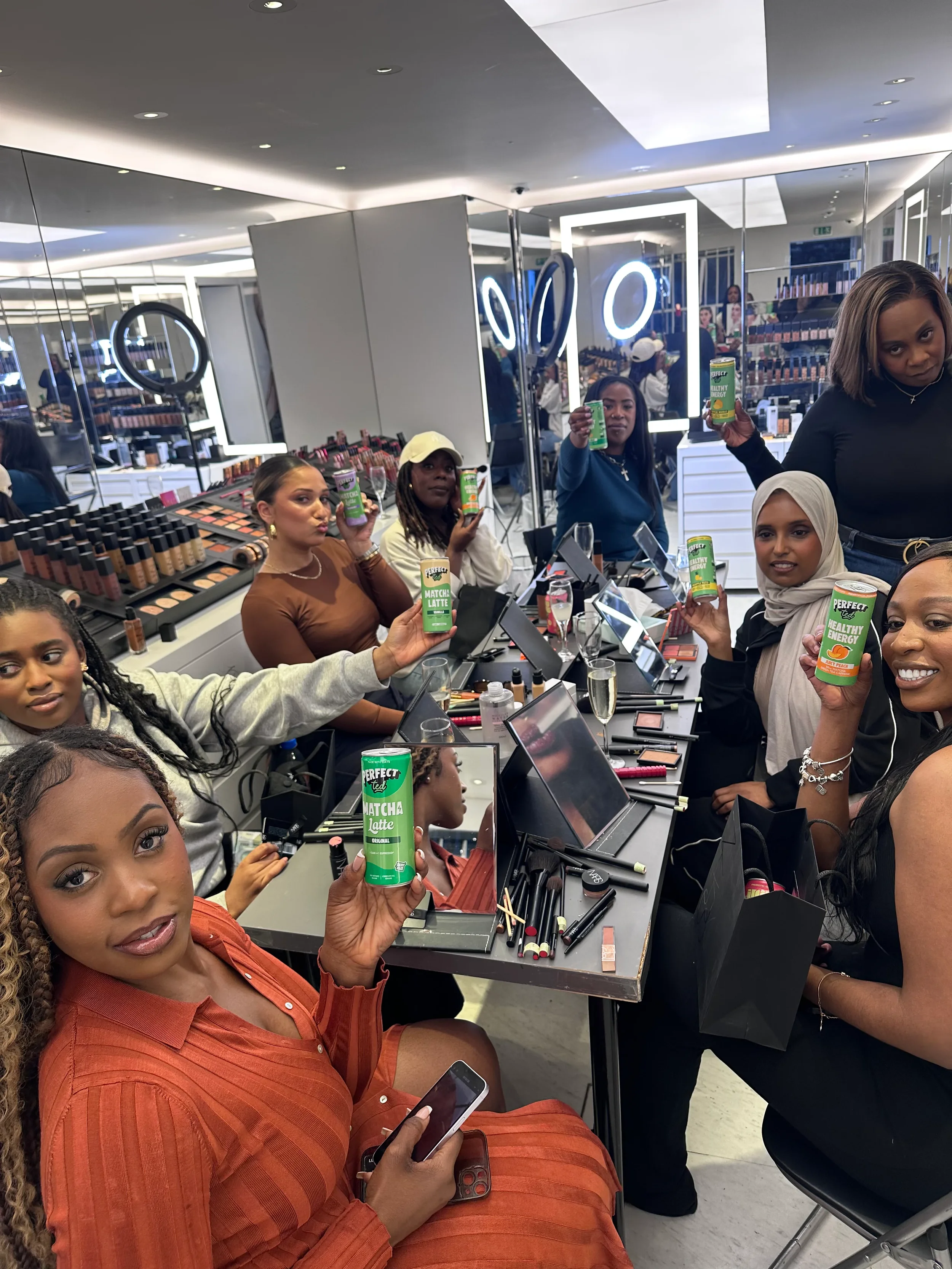 Group of women around a makeup counter holding cans of beverage, with makeup products and mirrors on the table, in a modern retail store.