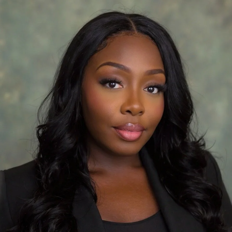 Close-up portrait of a woman with long wavy black hair, wearing a black top, neutral makeup, and a slight smile, against a blurred background.