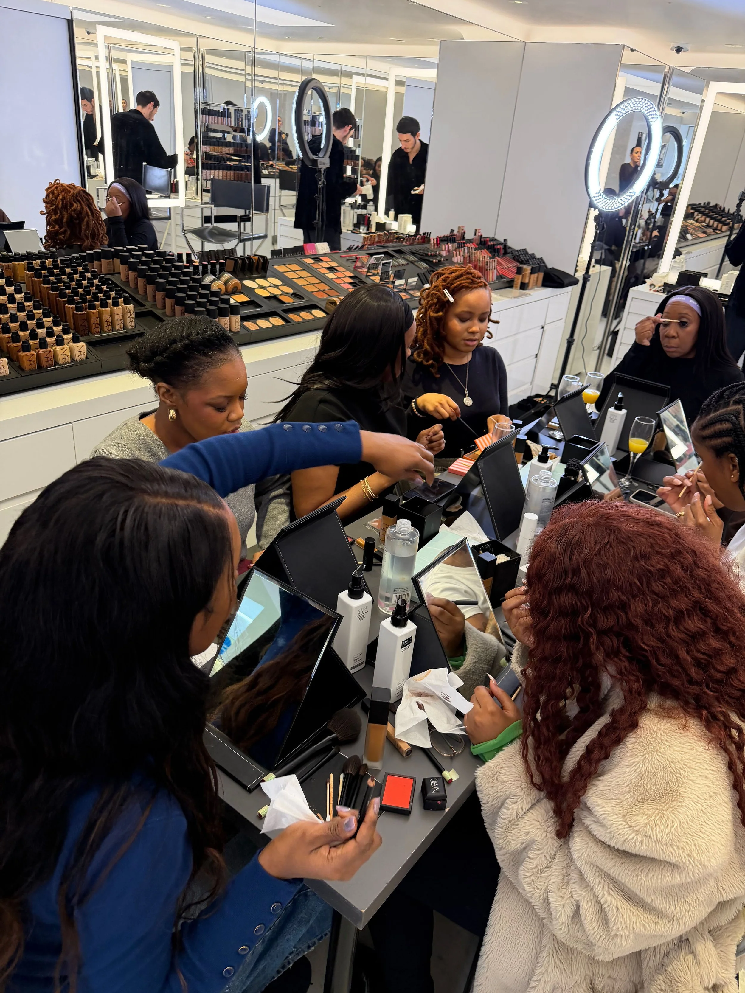 People shopping for makeup at a cosmetics counter in a store, with various makeup products displayed on the counter and mirrors with ring lights.