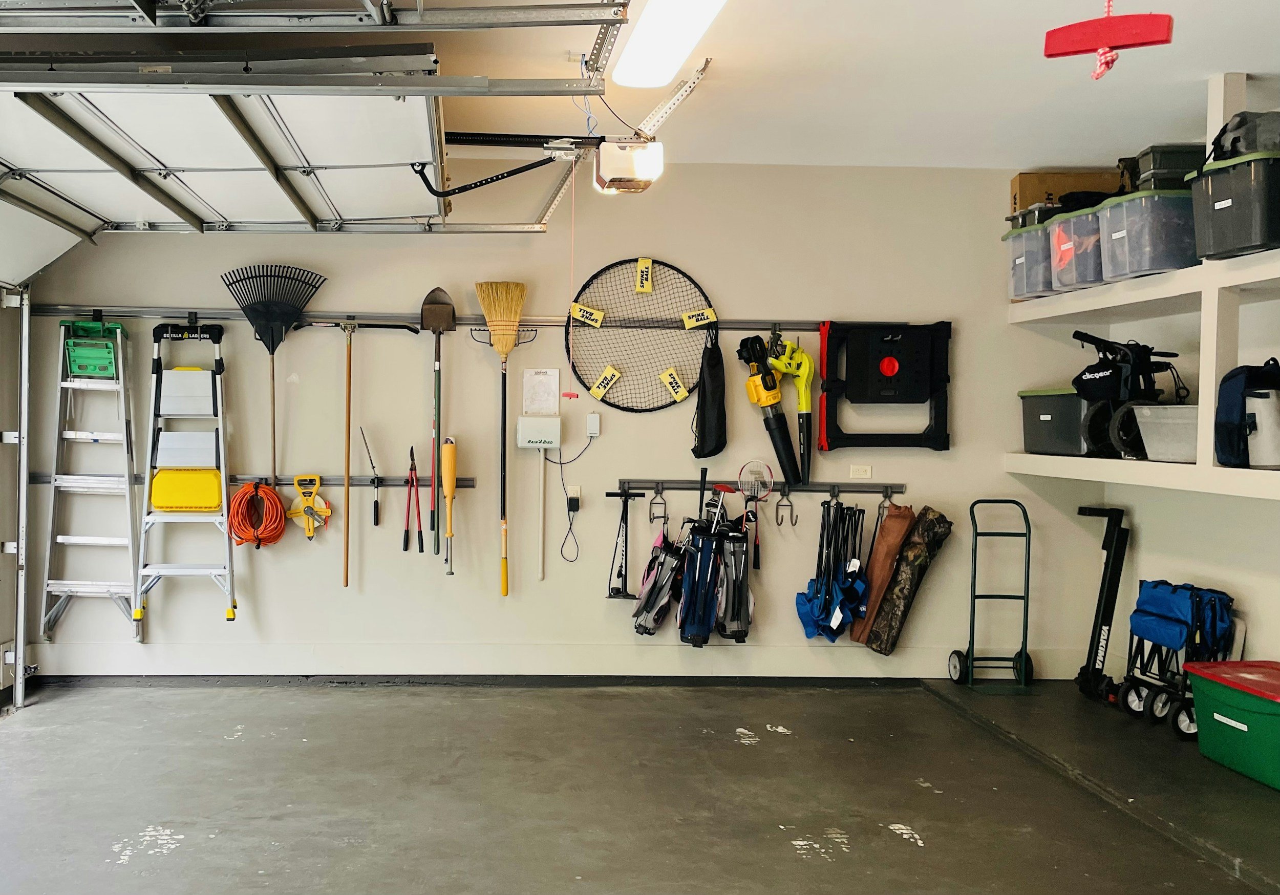 Garage wall with organized tools and storage items, including ladders, rakes, shovels, a wheelbarrow, and storage bins.